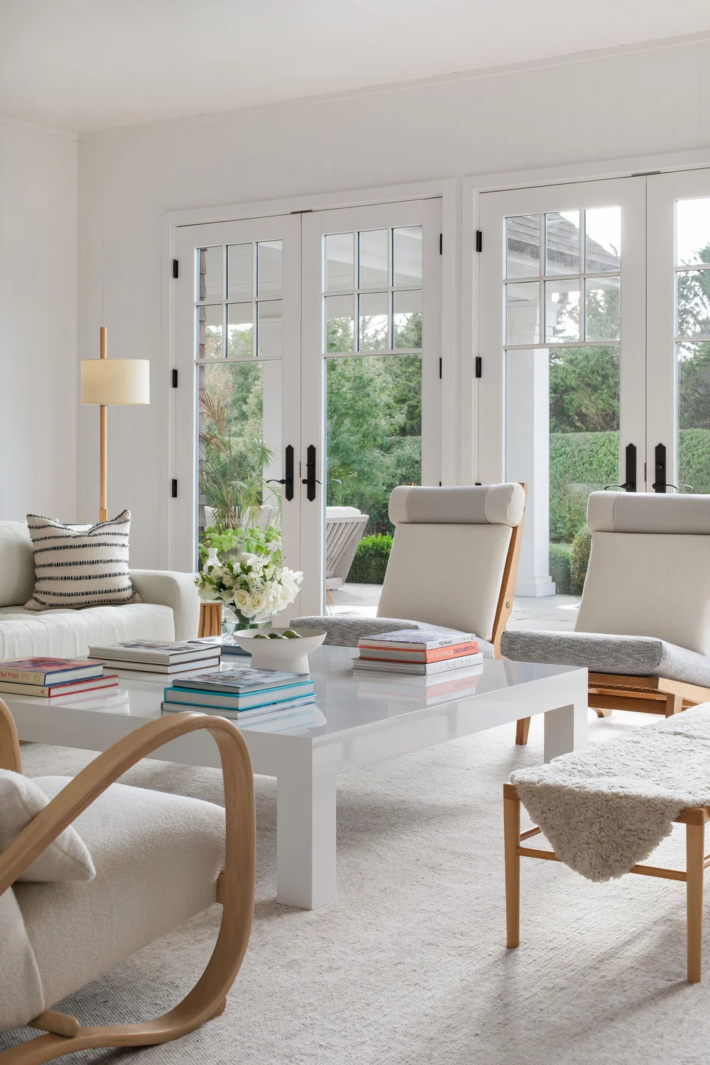 Bright living room with white furniture and a large white coffee table; glass French doors open to a green backyard; books and a flower vase decorate the table.