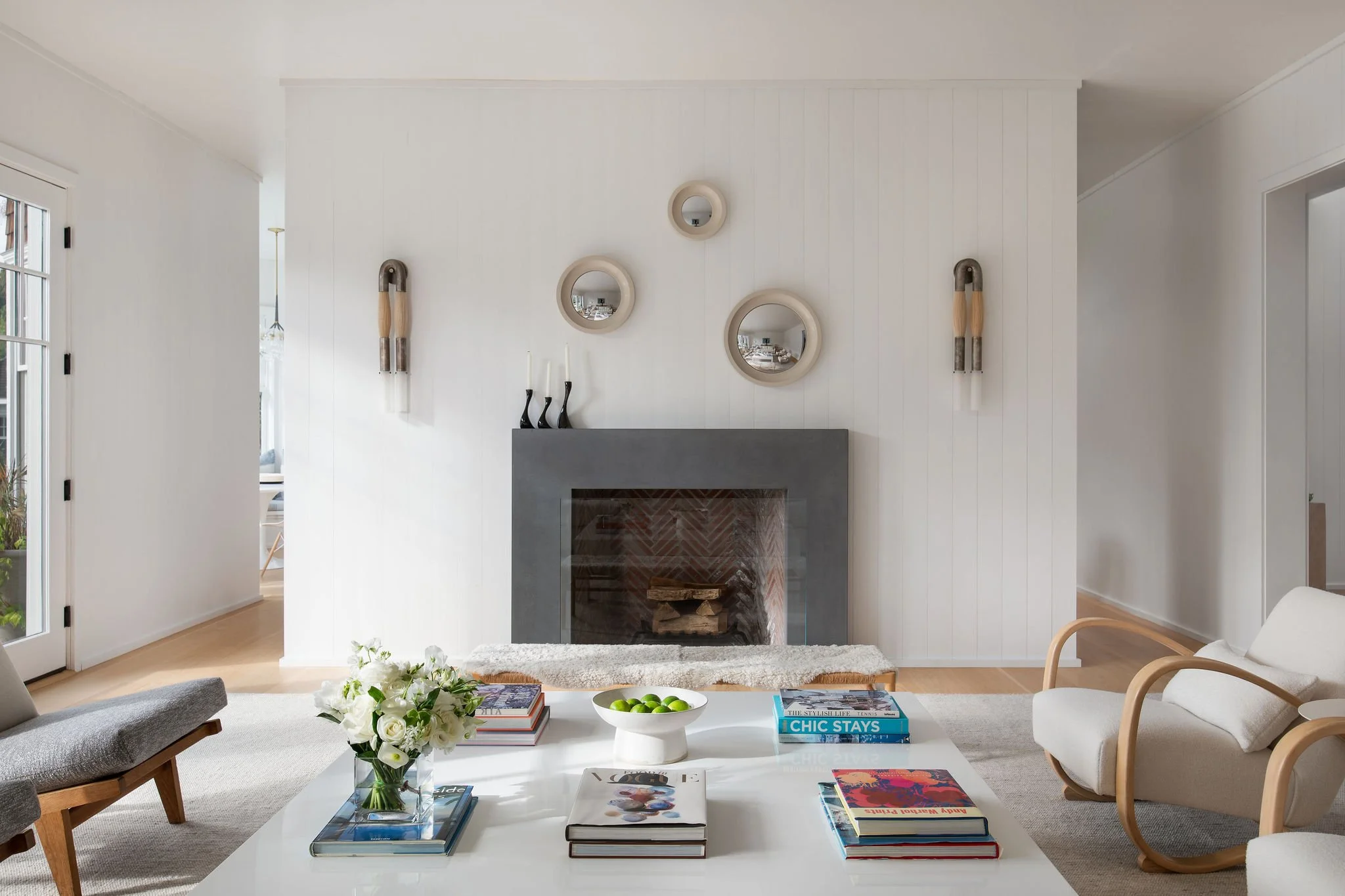 Bright living room with a white paneled wall, round mirrors above a fireplace, and a glass coffee table stacked with books and a vase of white flowers.