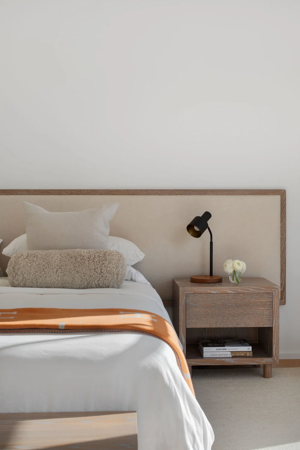 Bedroom with a beige headboard, white bedding, a textured tan throw, and an orange-accent blanket at the foot of the bed along with a nightstand.