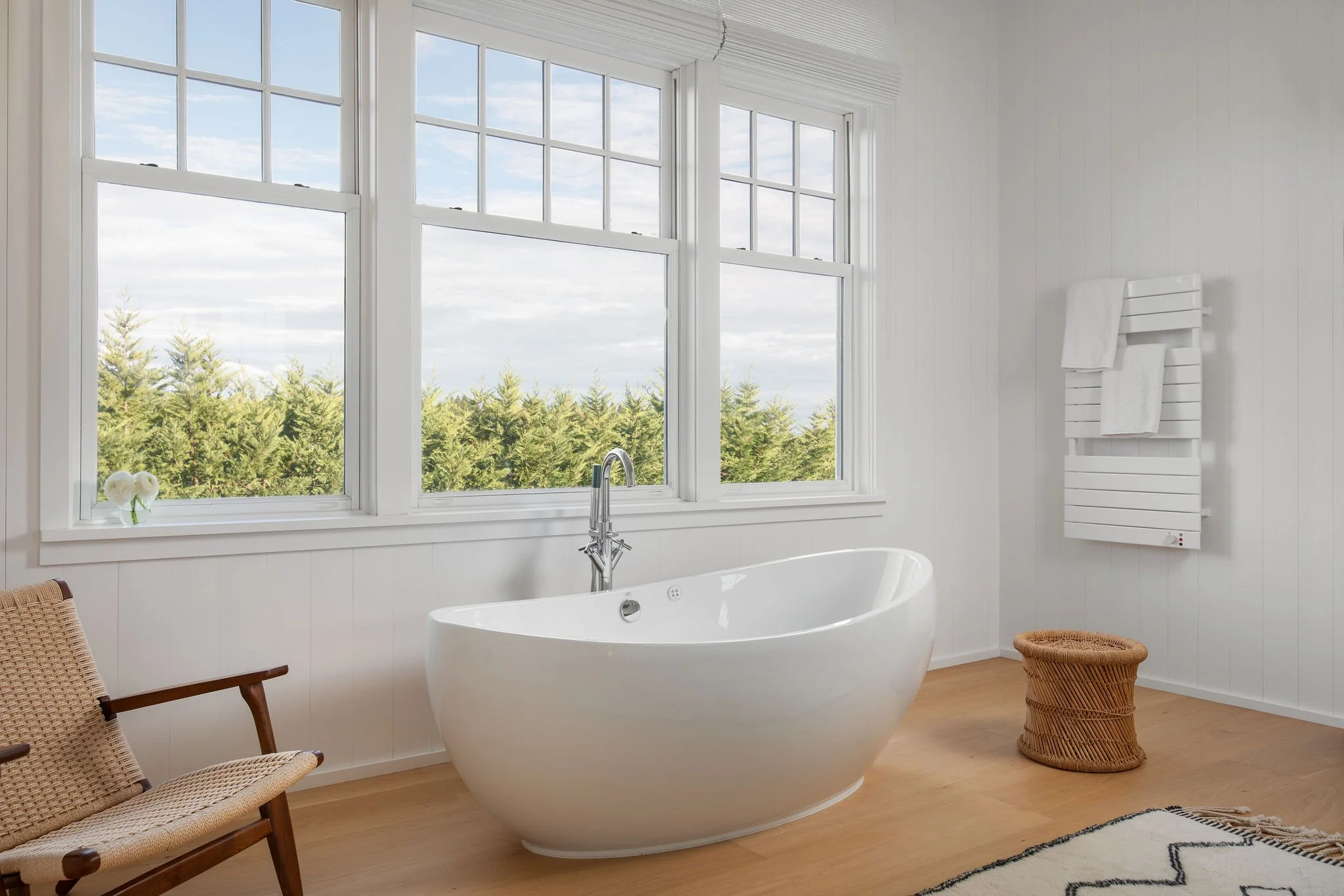 Bright white bathroom with a freestanding oval tub under large multi-pane windows overlooking trees outside.