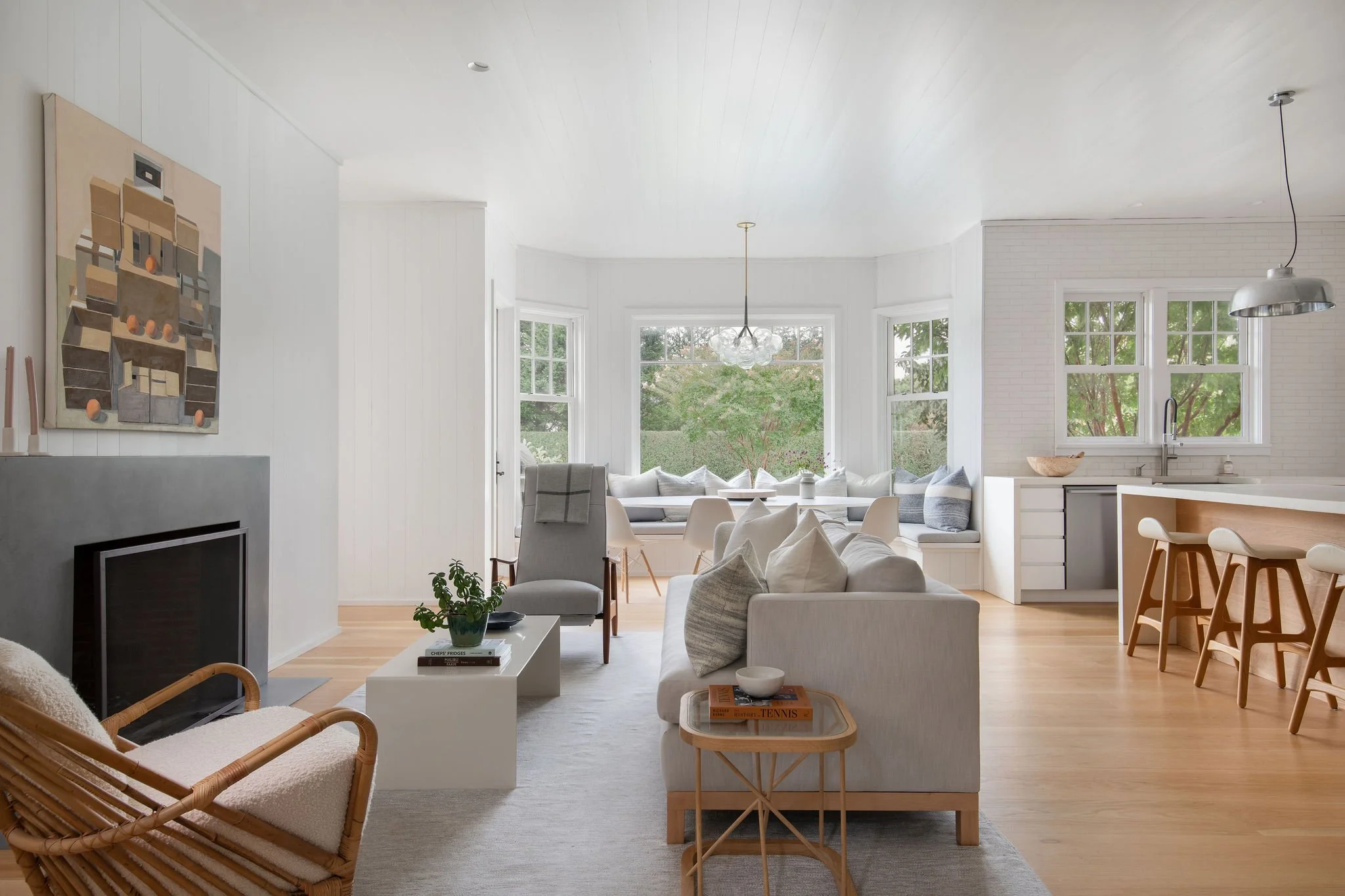Bright open living area with white walls, light wooden floors, large bay windows, and a kitchen island with stools.