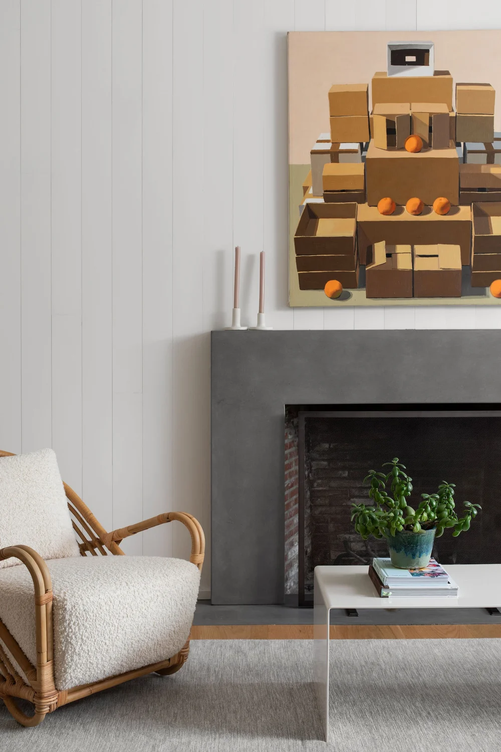 Living room corner with a rattan chair, gray fireplace, and a large abstract painting of stacked boxes above the mantel. A white coffee table with a potted plant sits in front, on a light rug.