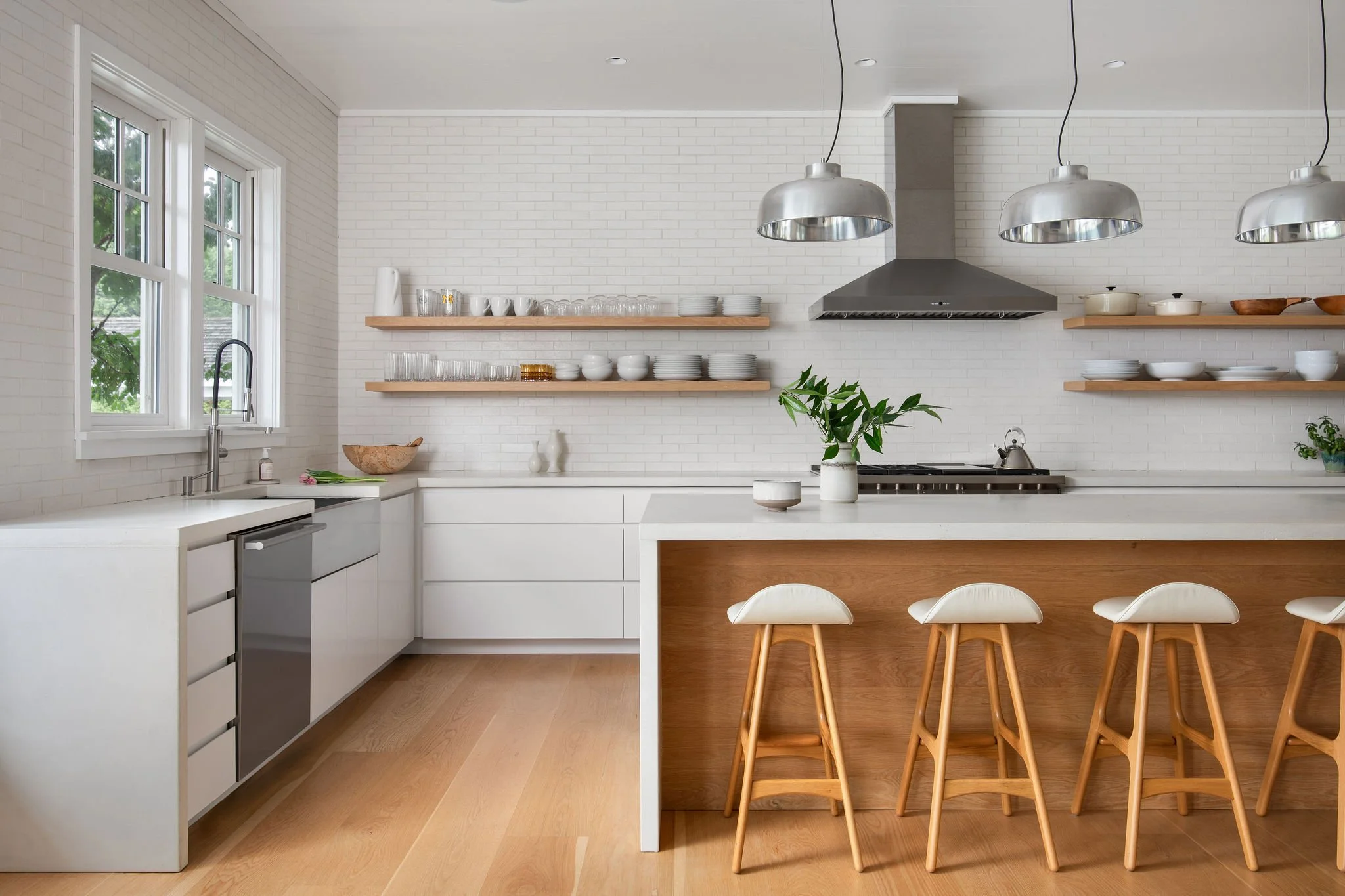 Bright white kitchen with subway tile backsplash, floating wooden shelves, and stainless pendant lights over a white island with wooden base and five bar stools at the counter.