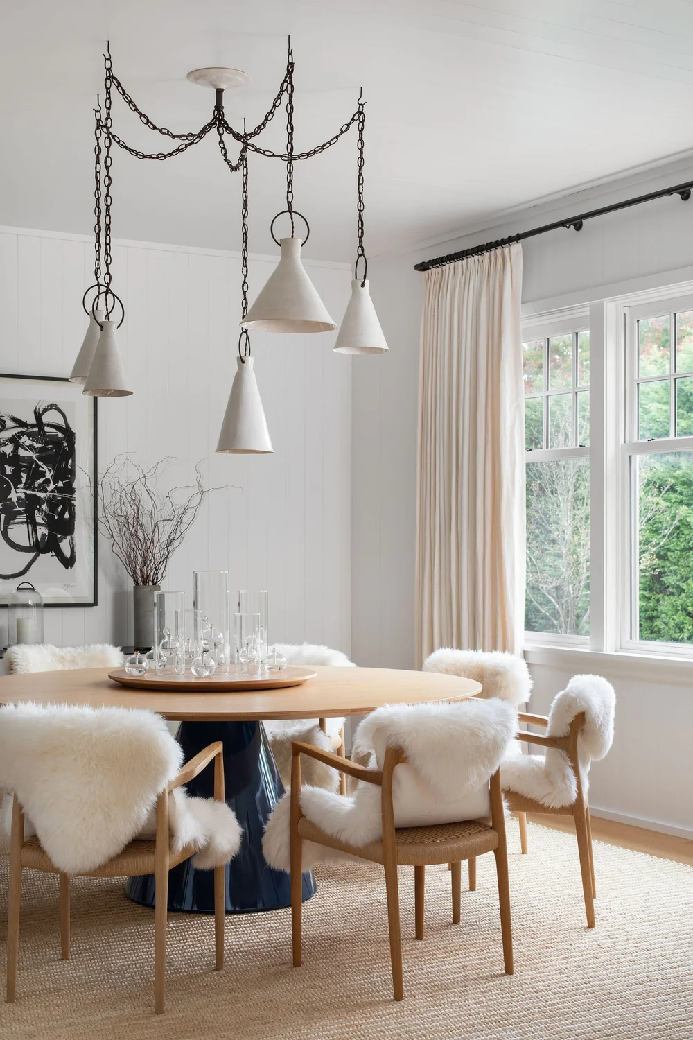 Round wooden dining table with blue pedestal base, surrounded by cream fur-covered chairs in a bright, minimal dining room.