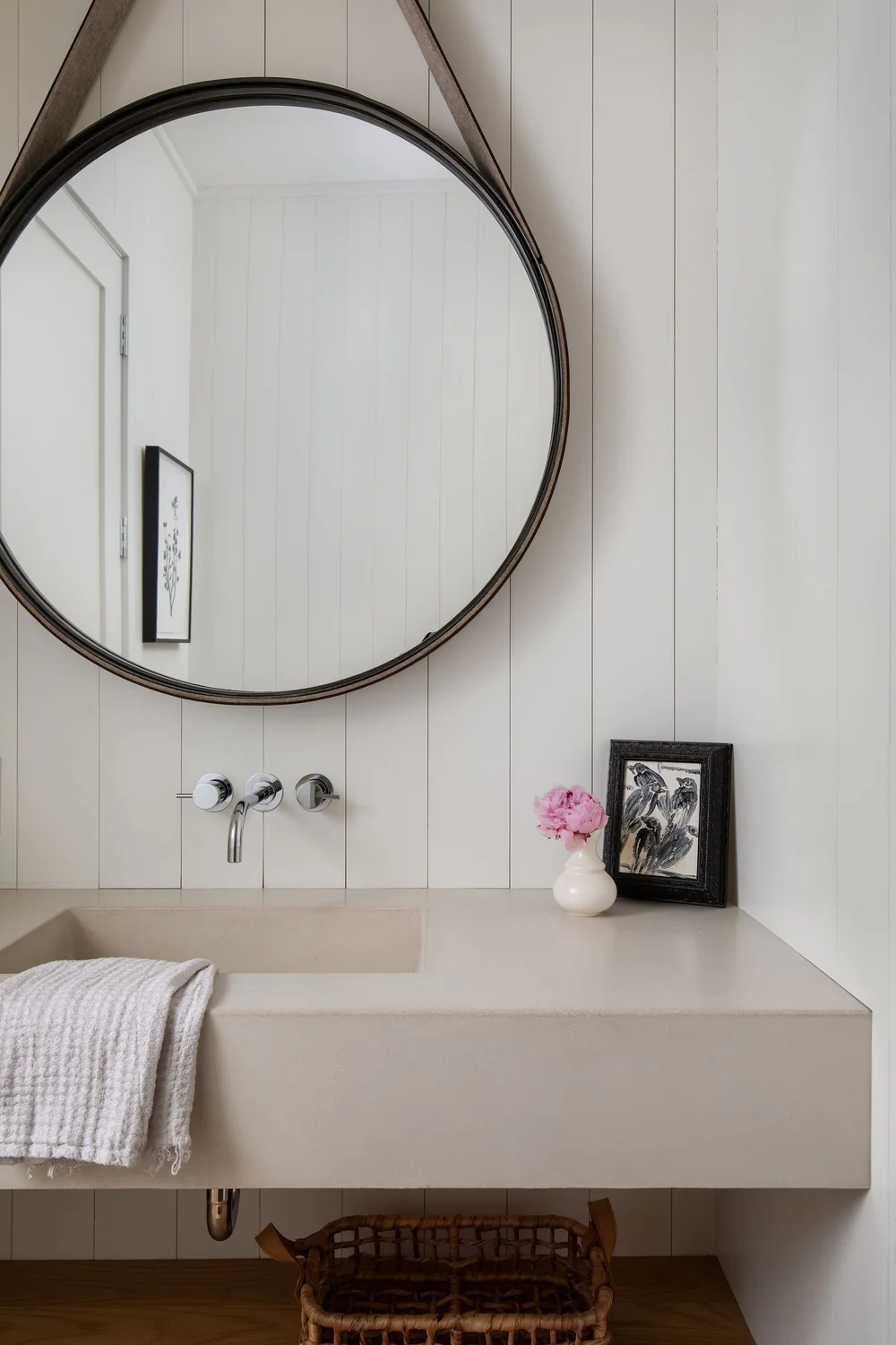 Round mirror above a beige countertop sink with wall faucets, a gray towel draped over the edge, and a small white vase with pink flowers beside a framed black-and-white art piece.