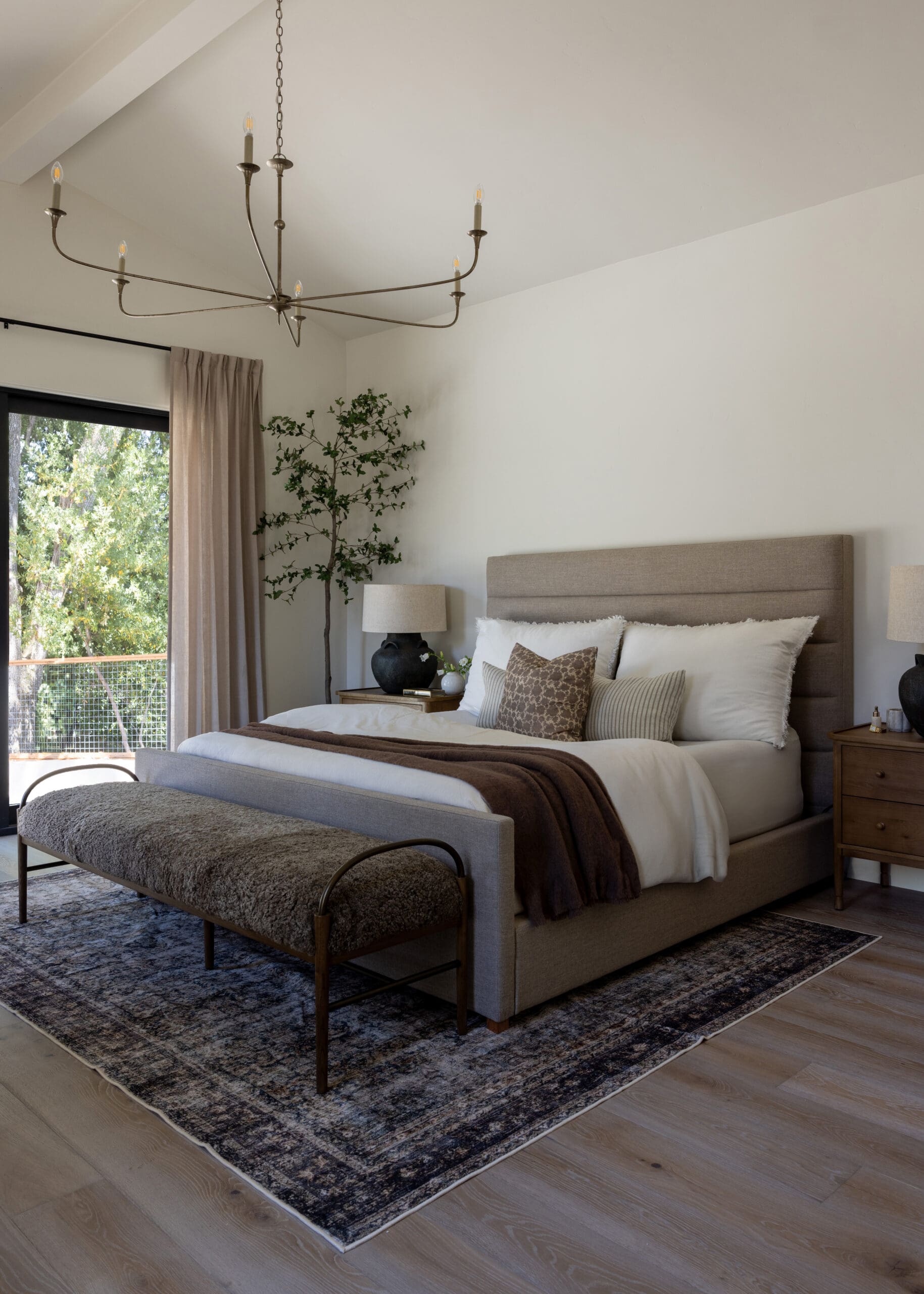 Neutral-toned bedroom with a beige upholstered bed, matching nightstands, and a four-armed metal chandelier hanging from the ceiling.
