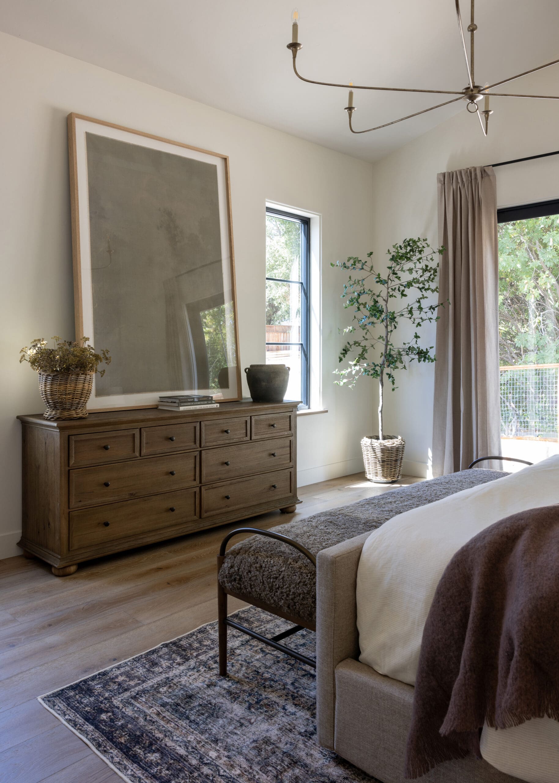 Bedroom corner with a wooden dresser, a large leaning framed artwork, and a basket with dried flowers on top. A tall potted tree stands by the window with sheer curtains.