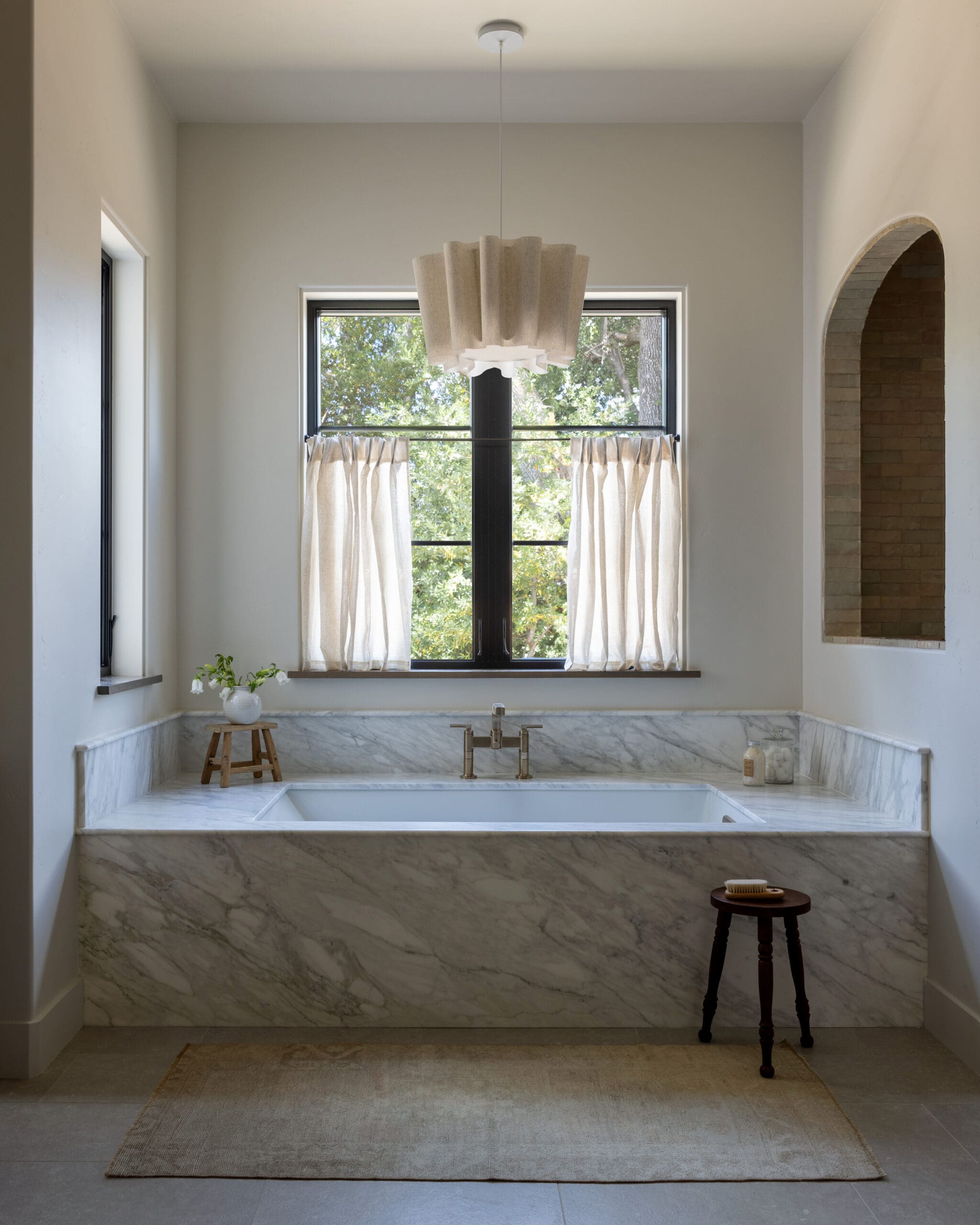 Marble bathroom with a built-in tub, window with sheer curtains, and a sculptural pendant light above the tub.