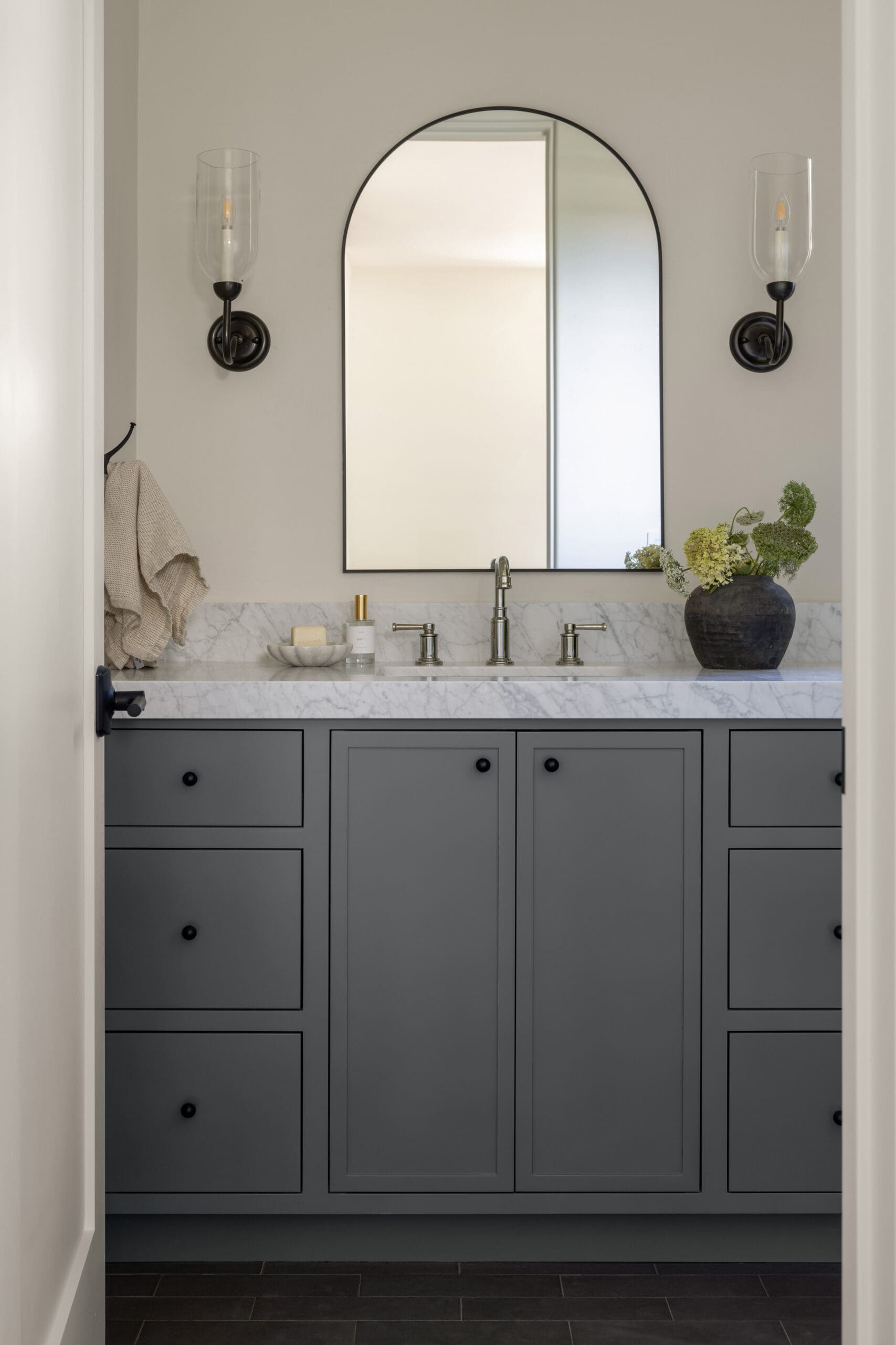 Gray bathroom vanity with marble countertop, arched mirror, and two black wall sconces flanking the mirror.