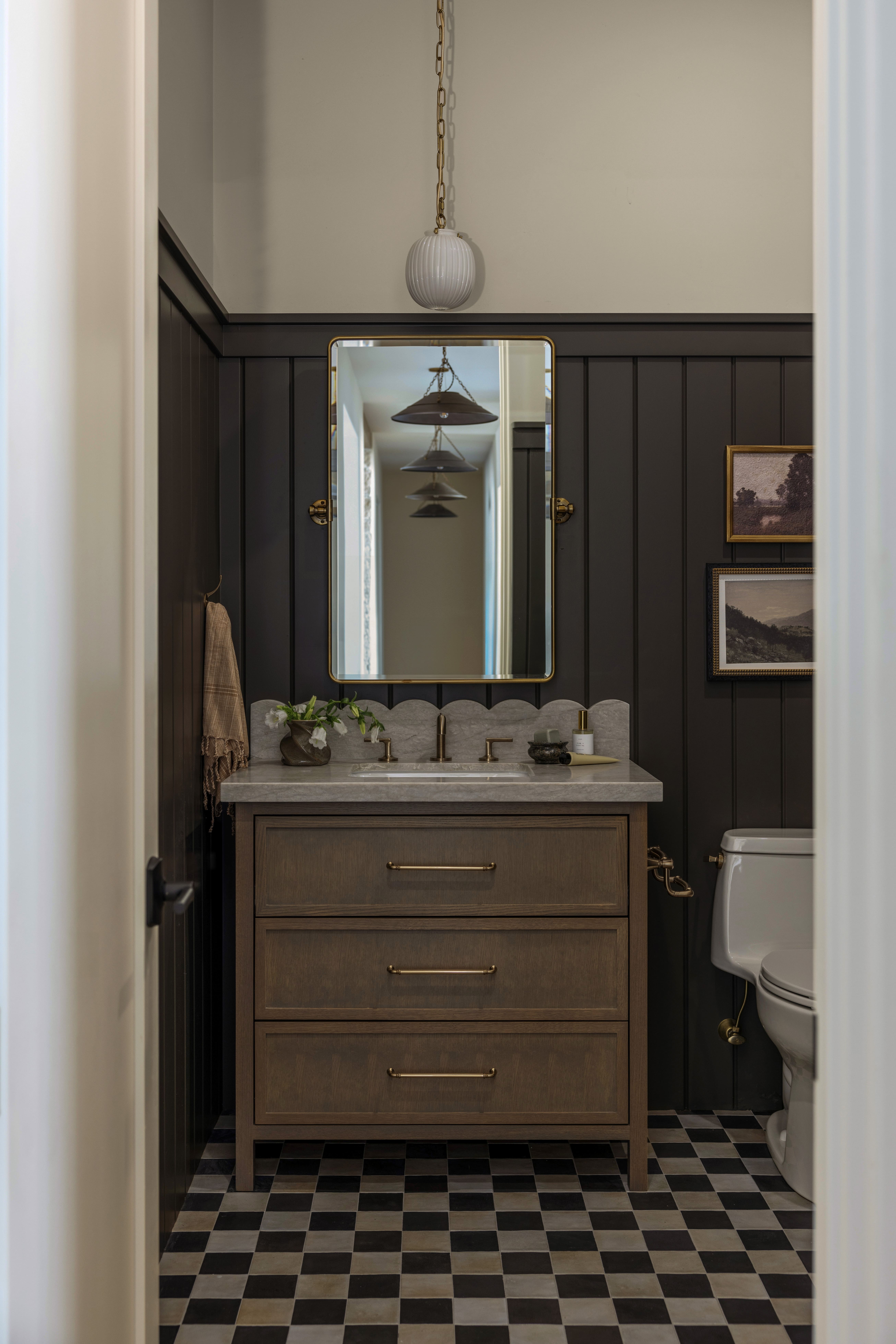 Dark-paneled bathroom vanity with a marble countertop, brass hardware, and a rectangular mirror above it, moody lighting.