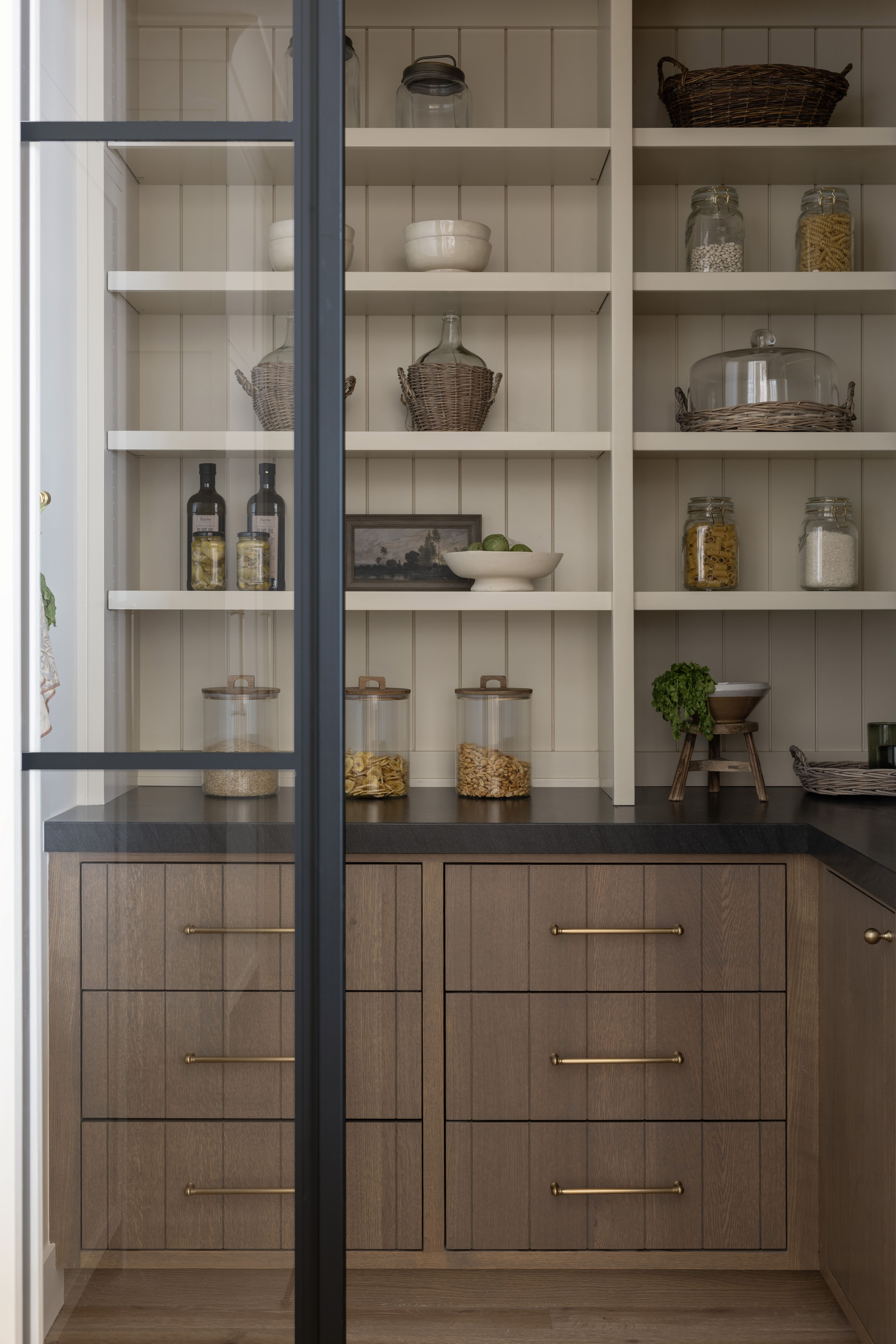 Open pantry with cream shelves holding glass jars, wicker baskets, ceramic bowls, and a small framed painting; glass cloche on the middle shelf.