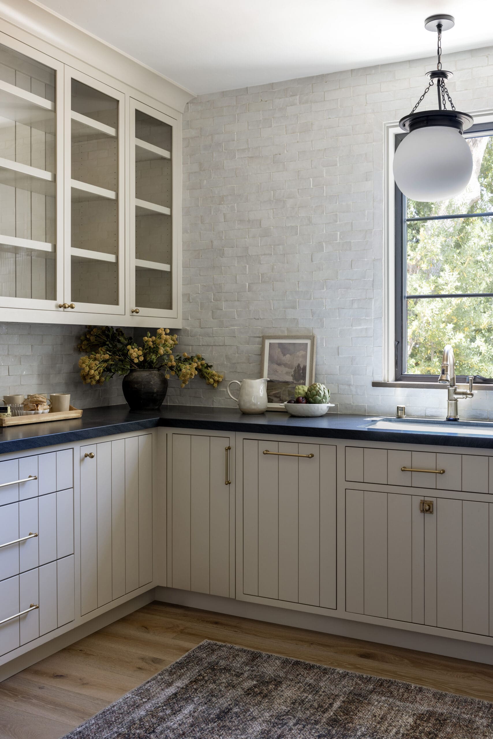 White kitchen corner with glass-front upper cabinets, gray brick backsplash, dark countertop, and a globe pendant light by a window over the sink lighted by natural light.