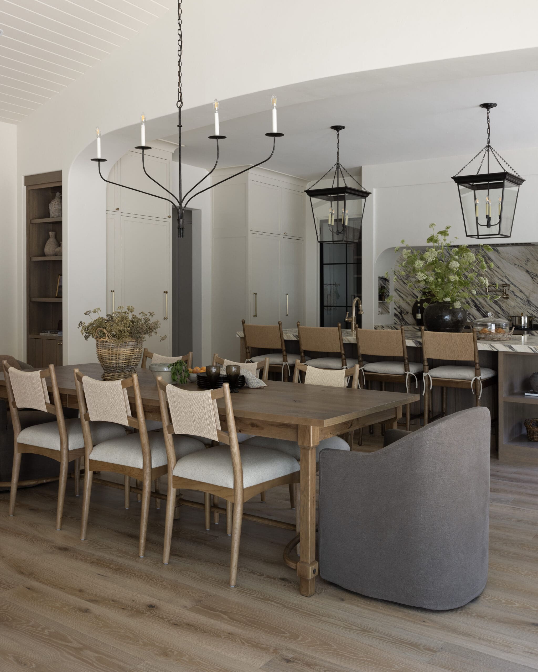 Open-concept kitchen and dining area featuring a long wooden table with beige upholstered chairs, white cabinets, and black metal chandeliers.