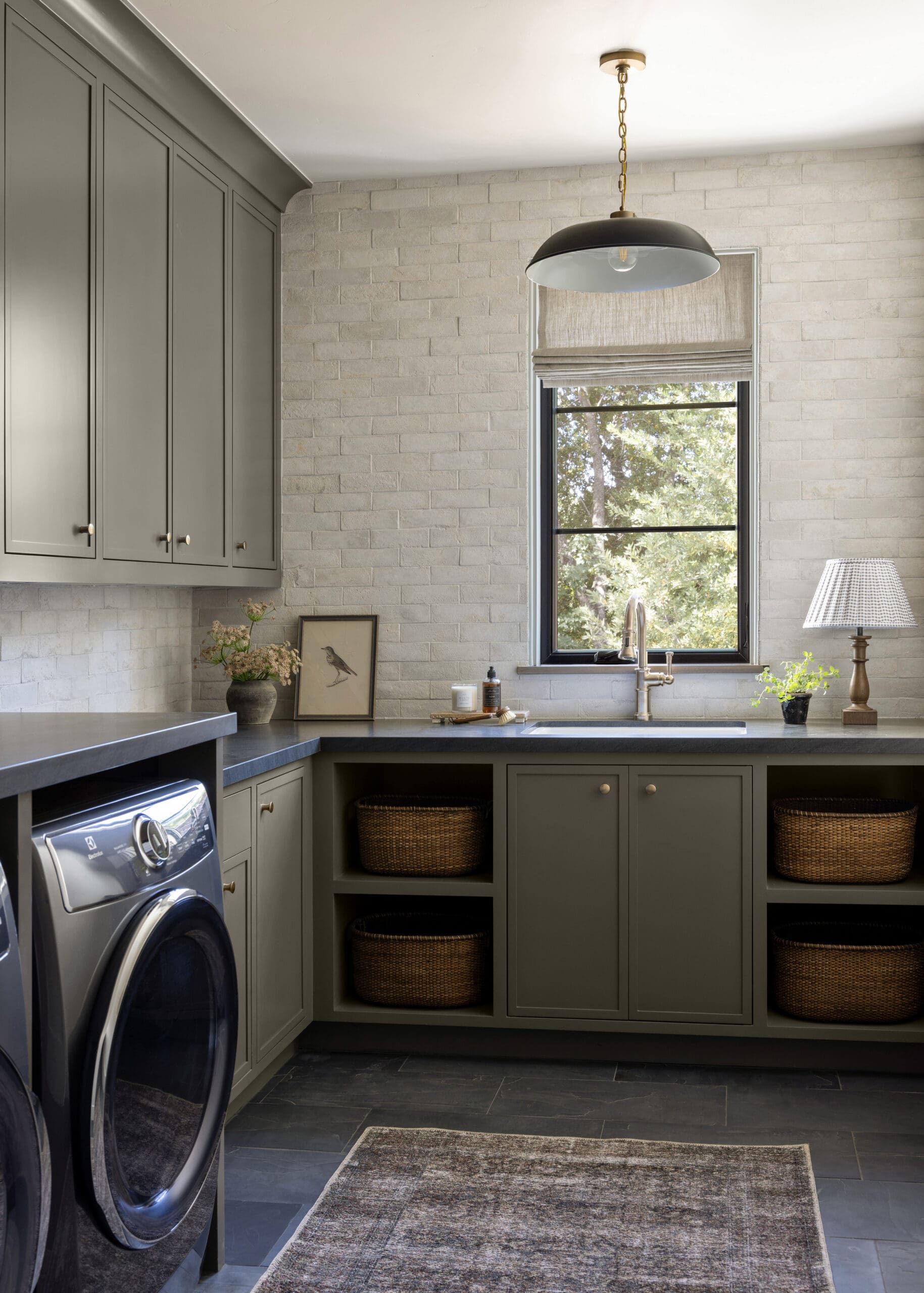 Laundry room with gray kitchen cabinets, brick wall, and a front-load washer and dryer under a dark countertop.