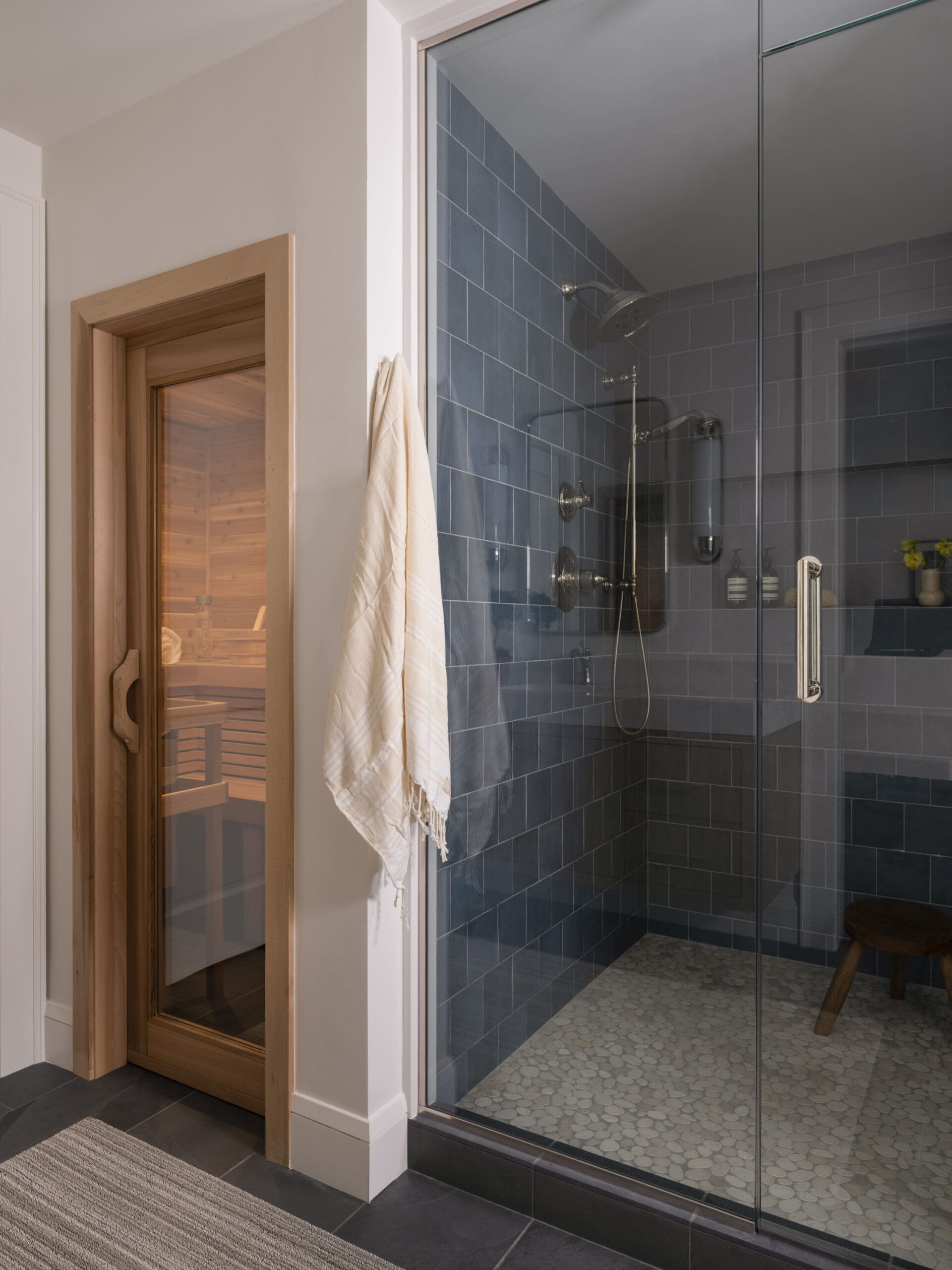 Glass‑enclosed shower in a modern bathroom with blue-gray tile walls, pebble floor, and a towel hanging on the door frame near a wooden sauna visible through the doorway.