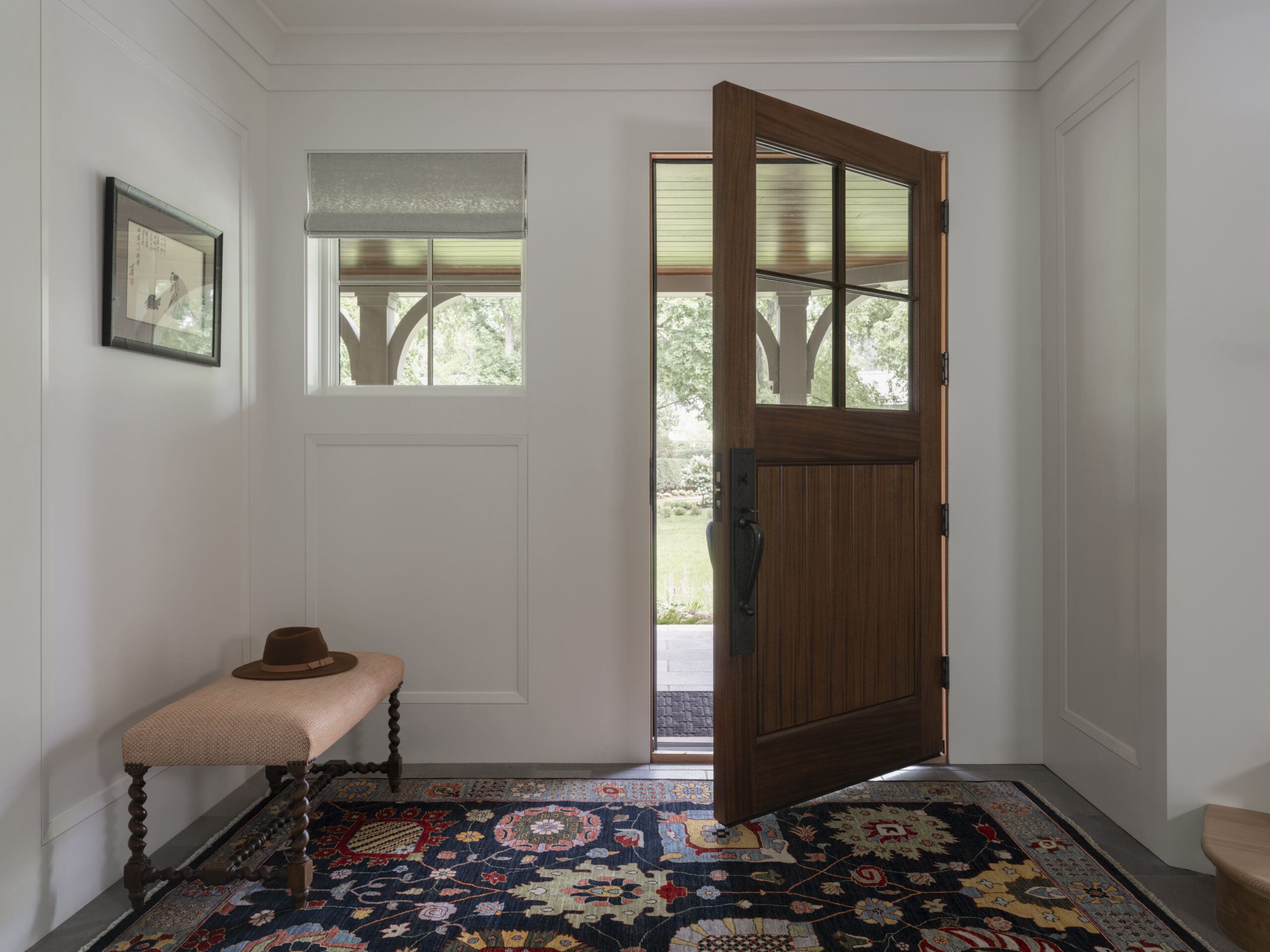 Open wooden front door leading outside, inside a white-paneled entryway with a patterned rug and a cushioned bench with a hat on it.