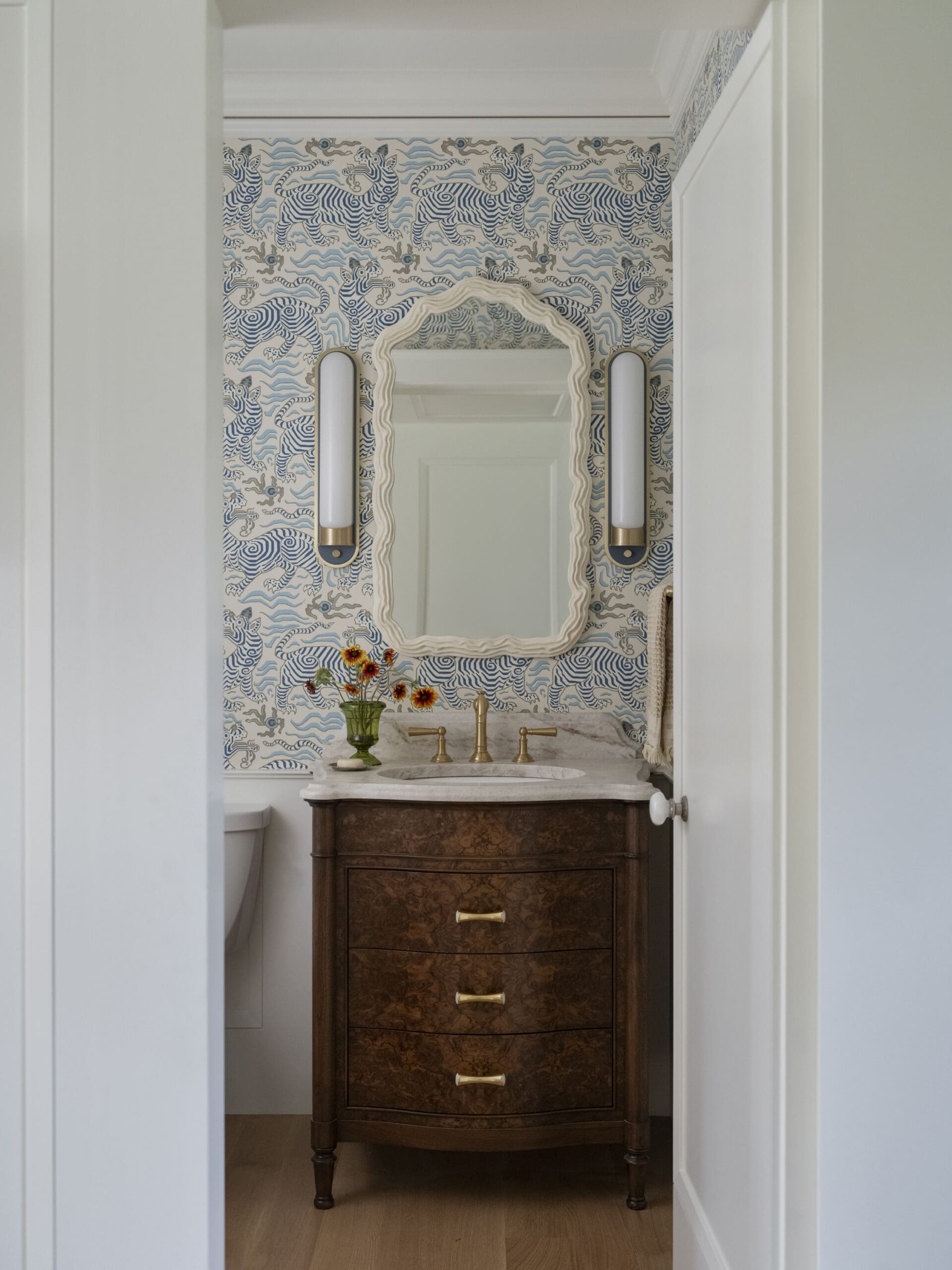 Bathroom features a dark wood vanity with marble top, brass faucet, wavy-framed mirror, blue zebra wallpaper, and a vase of flowers.