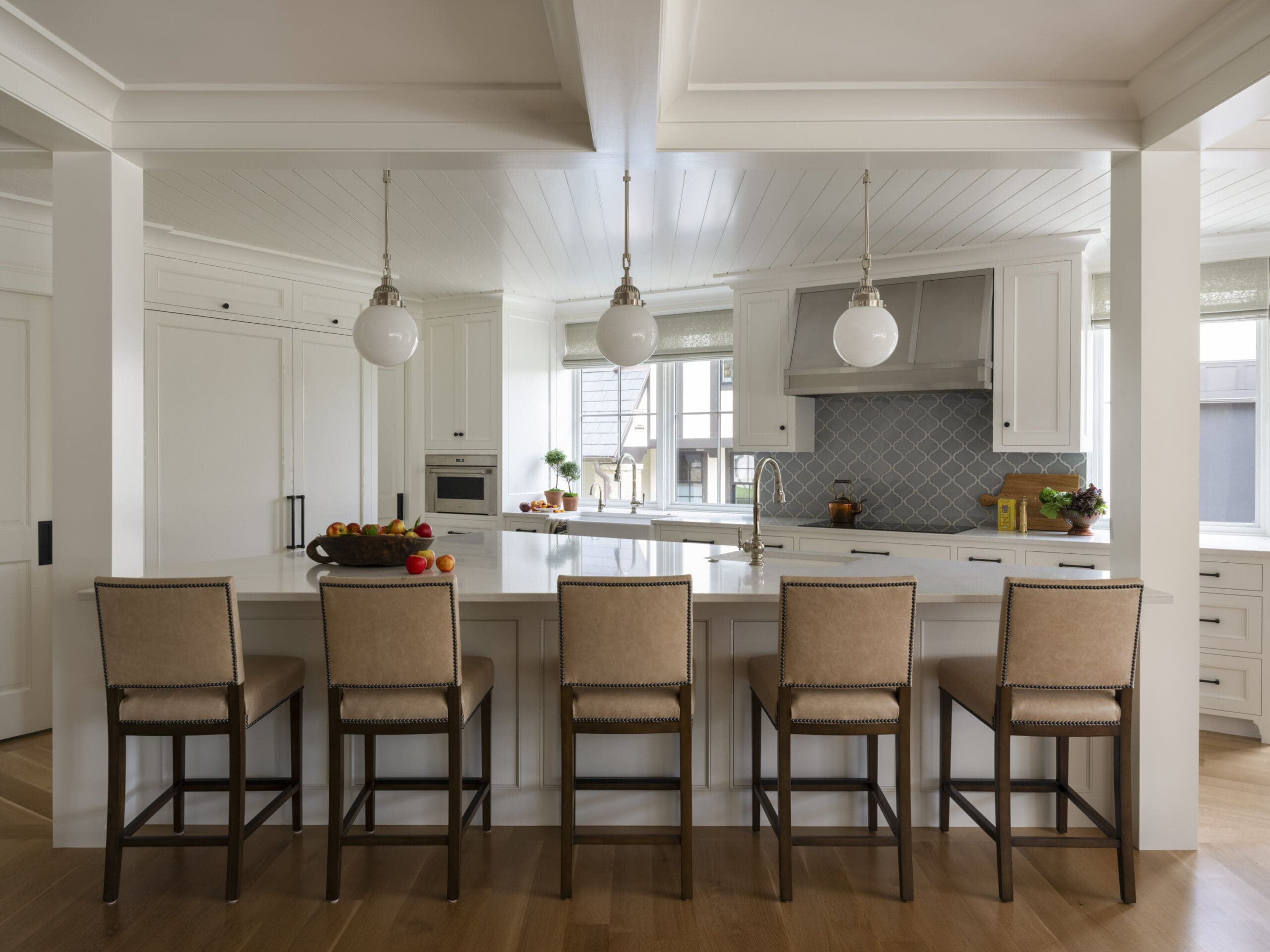 Bright white kitchen with a large island and five beige upholstered bar stools; pendant globes hang over the counter, overhead white cabinetry and a patterned backsplash near the stove.