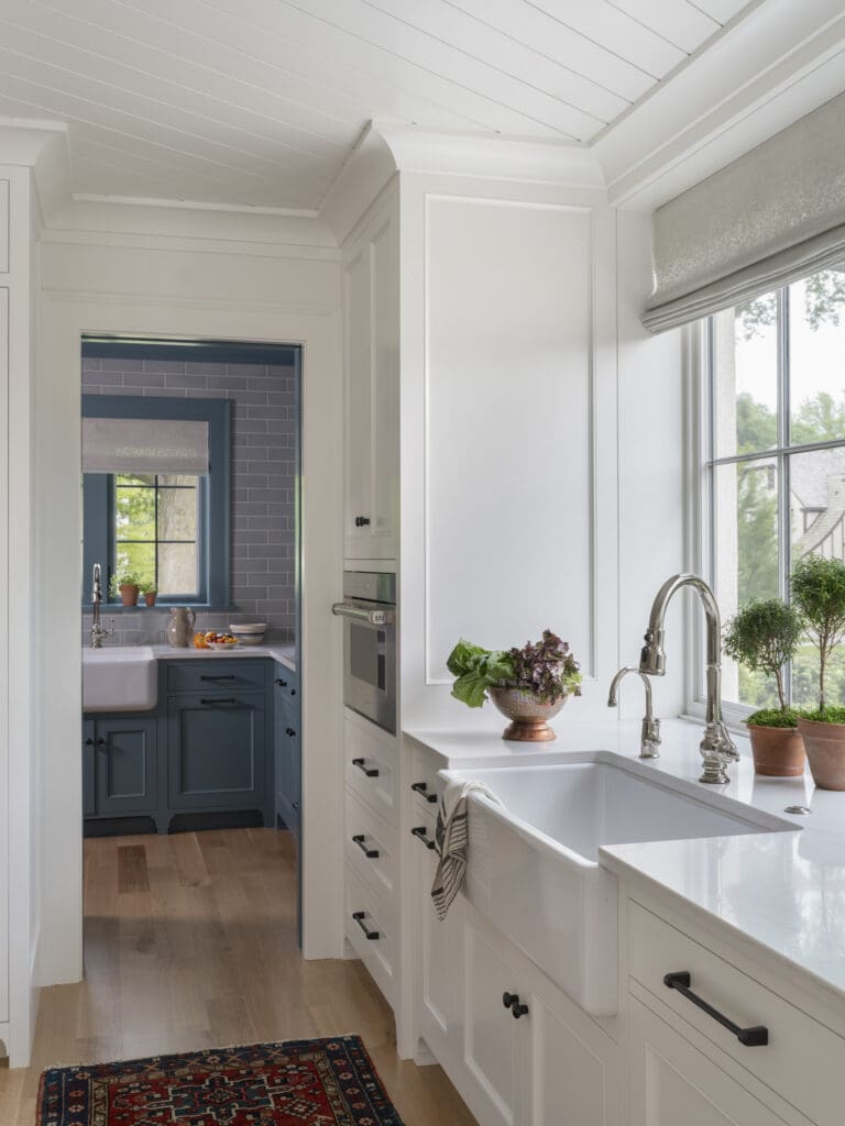 Bright white kitchen with large window, porcelain sink, and chrome taps; copper bowls with greens on the counter; view of a second, blue-toned kitchen through a doorway in the background.