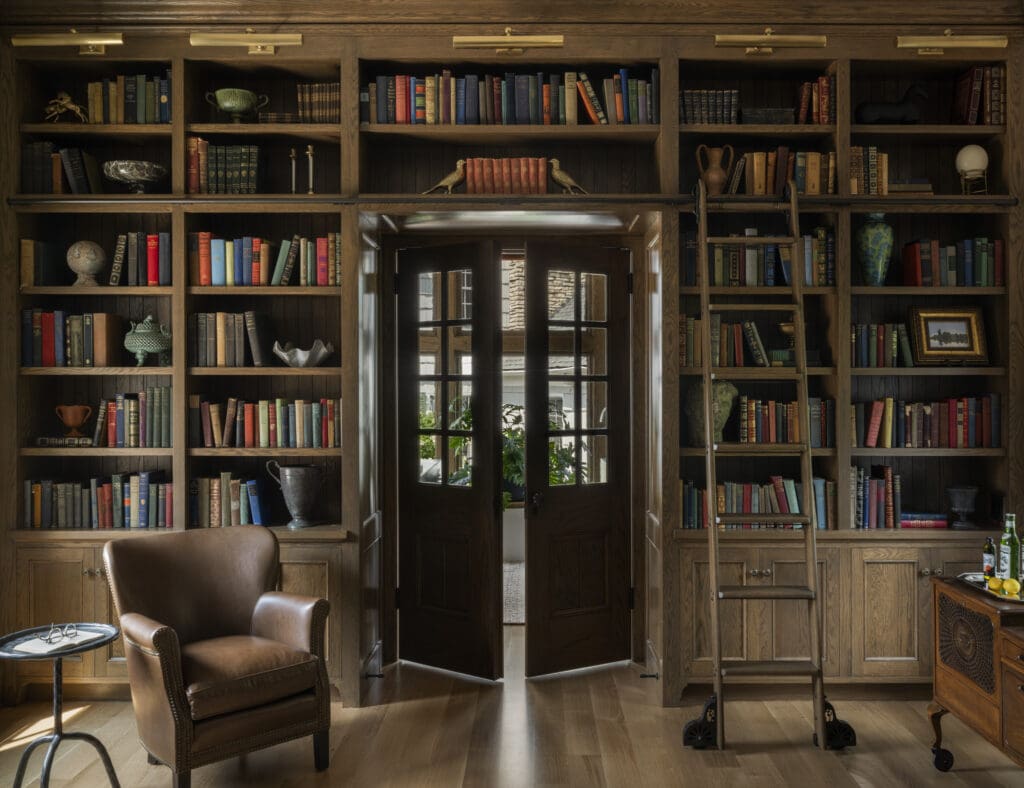 Cozy wooden library with tall built-in shelves, a rolling ladder, and a doorway opening to a sunlit room.