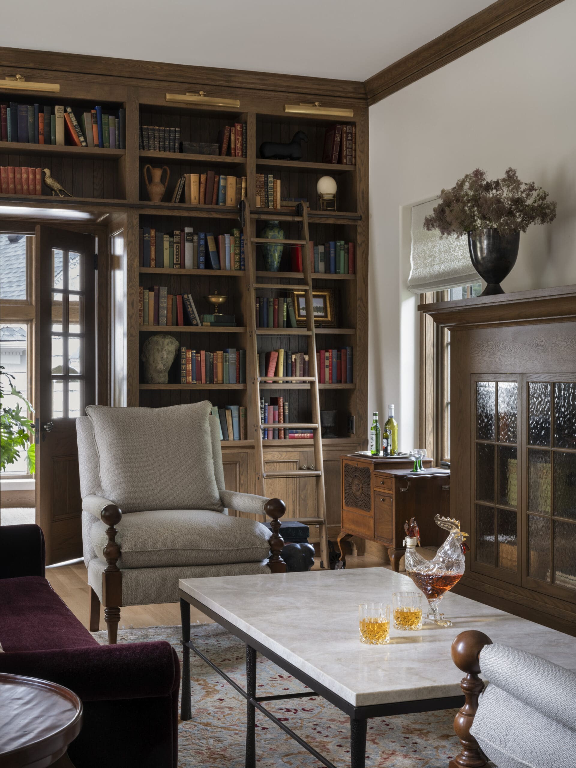 Cozy living room with tall wooden bookcases, a ladder, beige armchair, and a marble coffee table by a windowed wall.