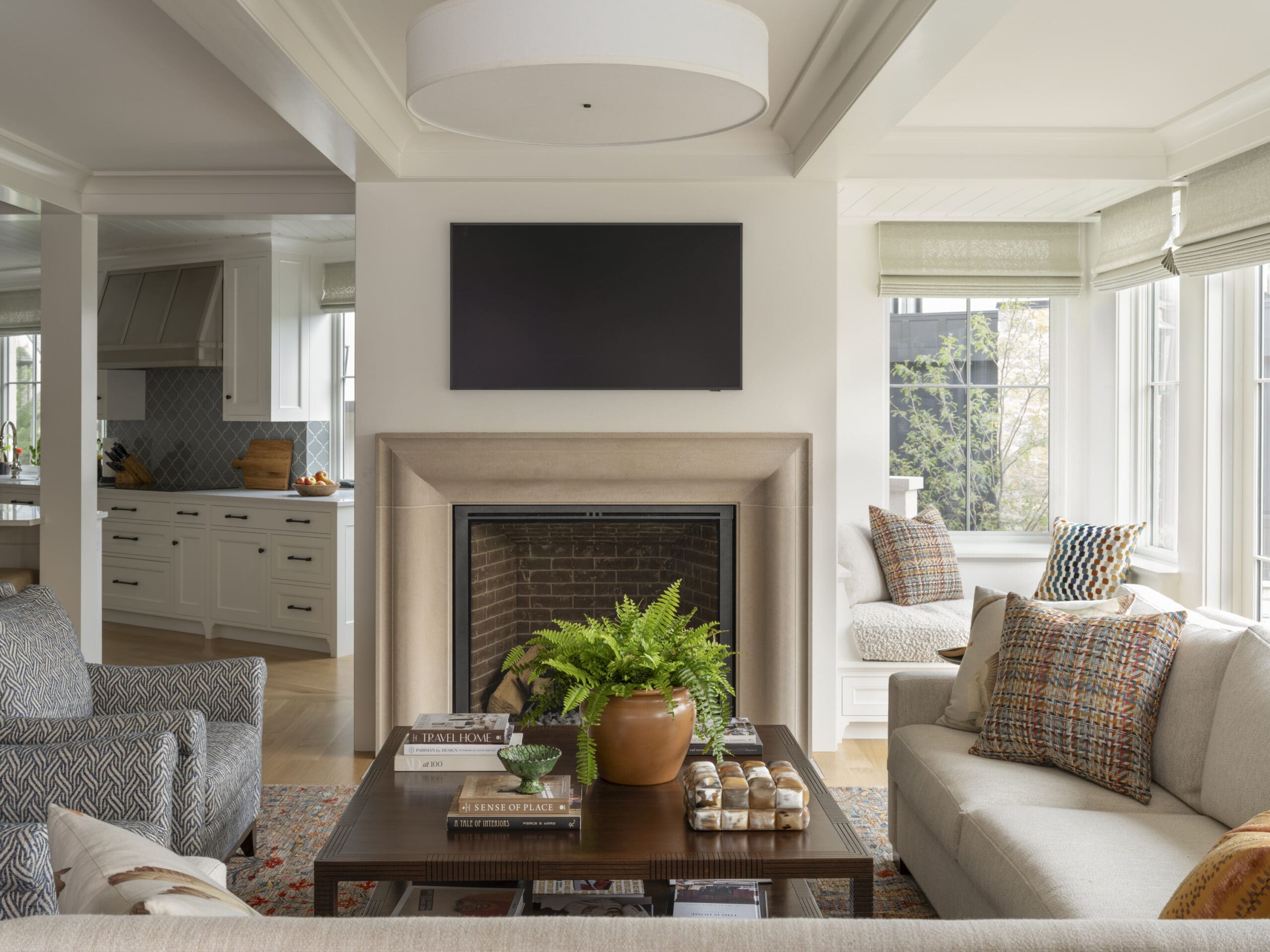 Living room with a beige fireplace, wall-mounted TV, and large windows with light curtains; blue patterned sofas nearby.