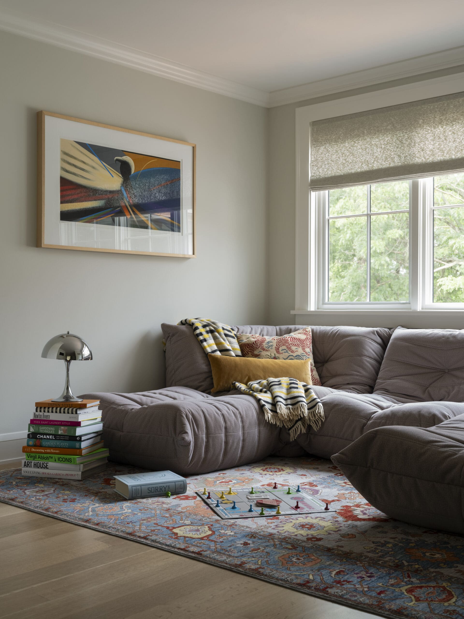 Cozy living room with a gray tufted sofa, colorful pillows, a striped blanket, abstract framed art, and a window with a textured blind.