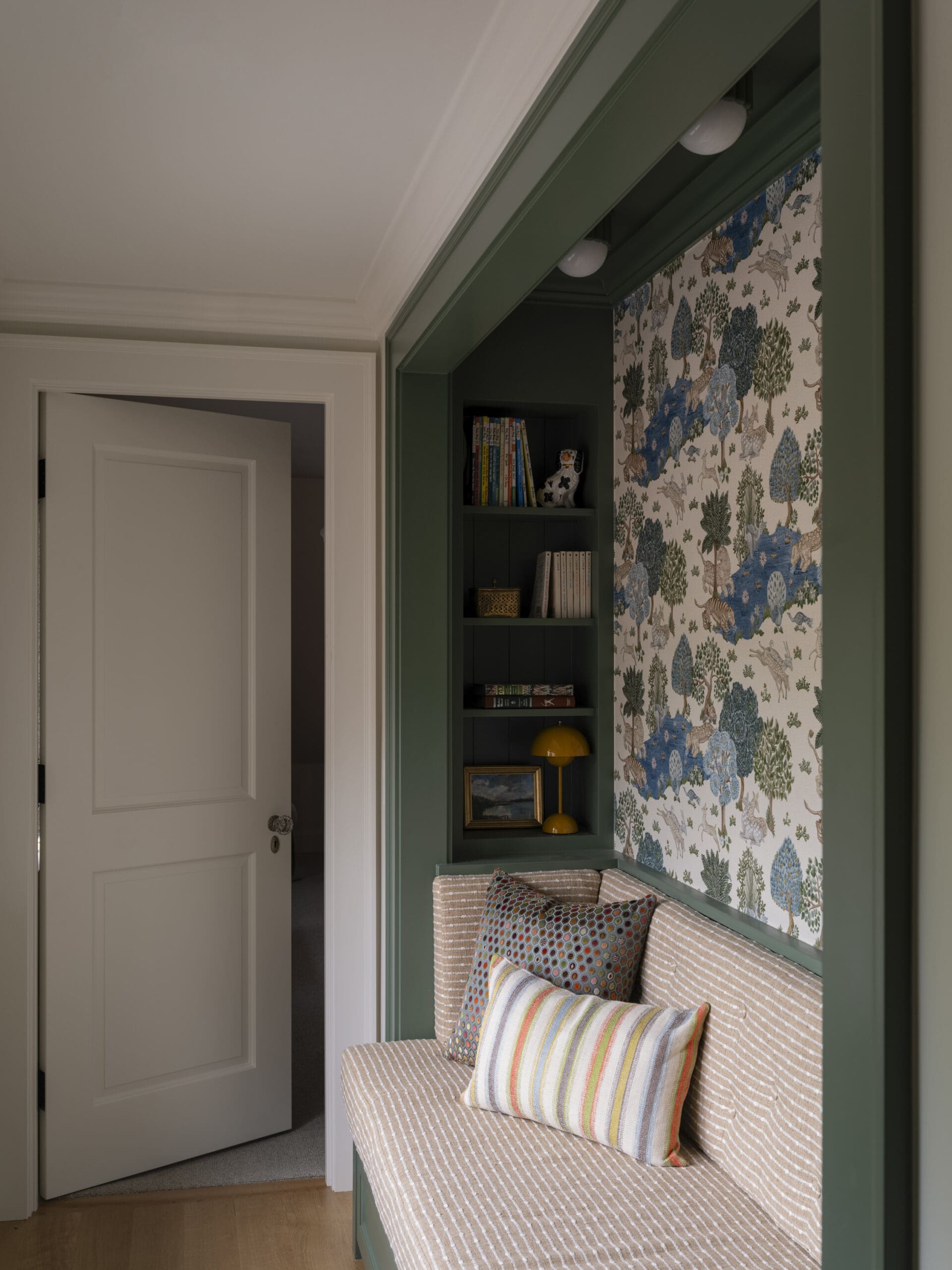 Built-in green alcove with shelves, books and a ceramic cow figurine, next to a beige striped sofa with colorful cushions, against botanical wallpaper.