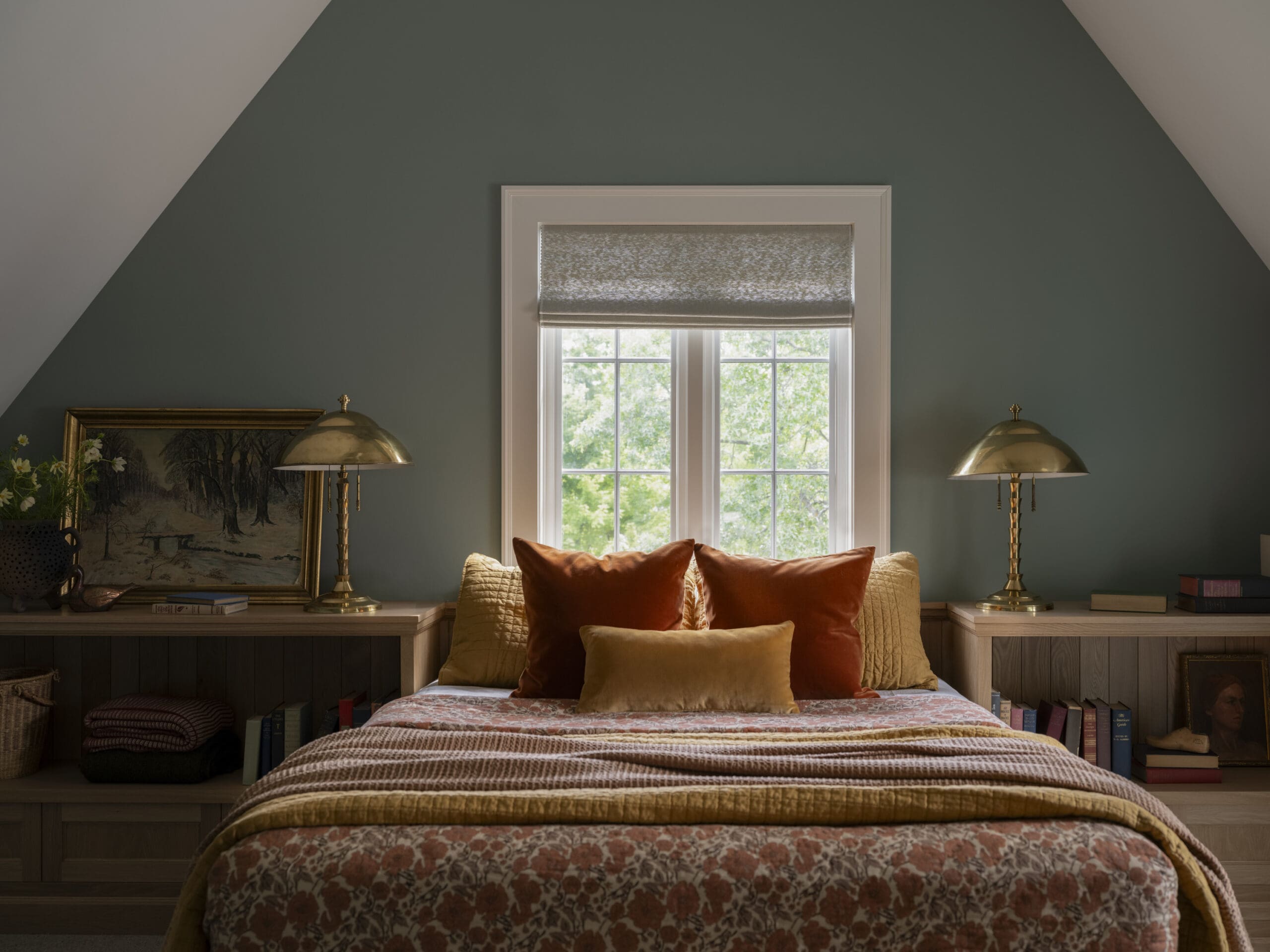 Cozy bedroom with a dark green wall, a window with a roman shade, and two brass lamps flanking a bed with orange and gold pillows.
