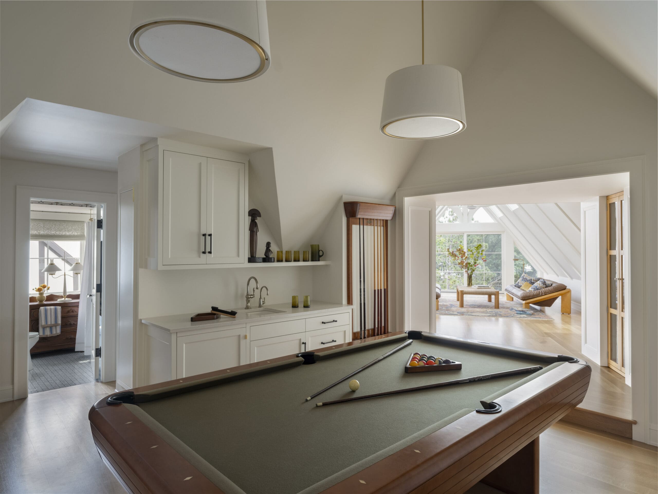 Open-plan interior showing a pool/billiards table in the foreground, white cabinetry to the left, and a bright living room visible through an doorway in back.