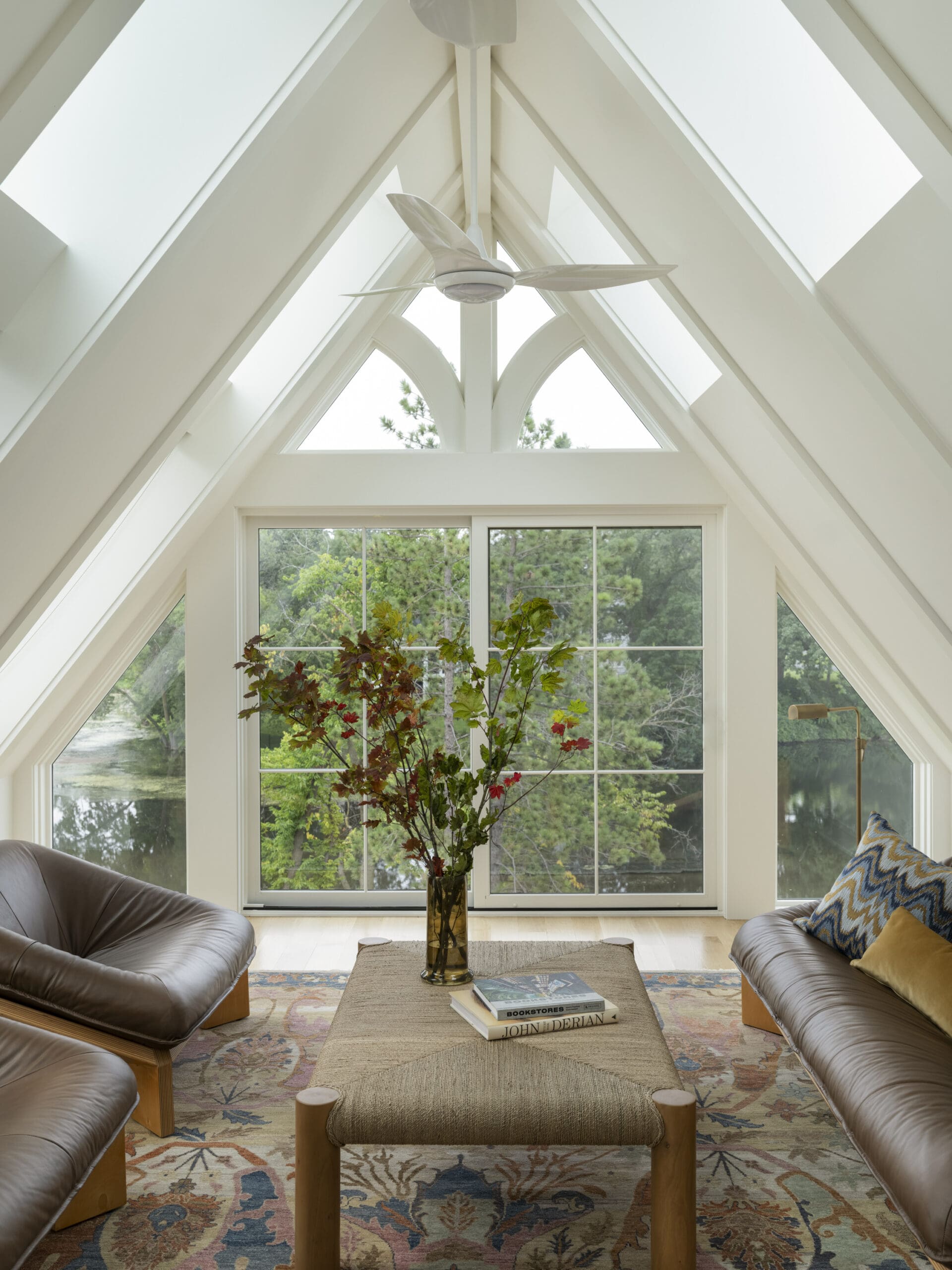Bright living room with a vaulted, triangular ceiling and large window wall; a tall vase with branches sits on a coffee table between two leather chairs.