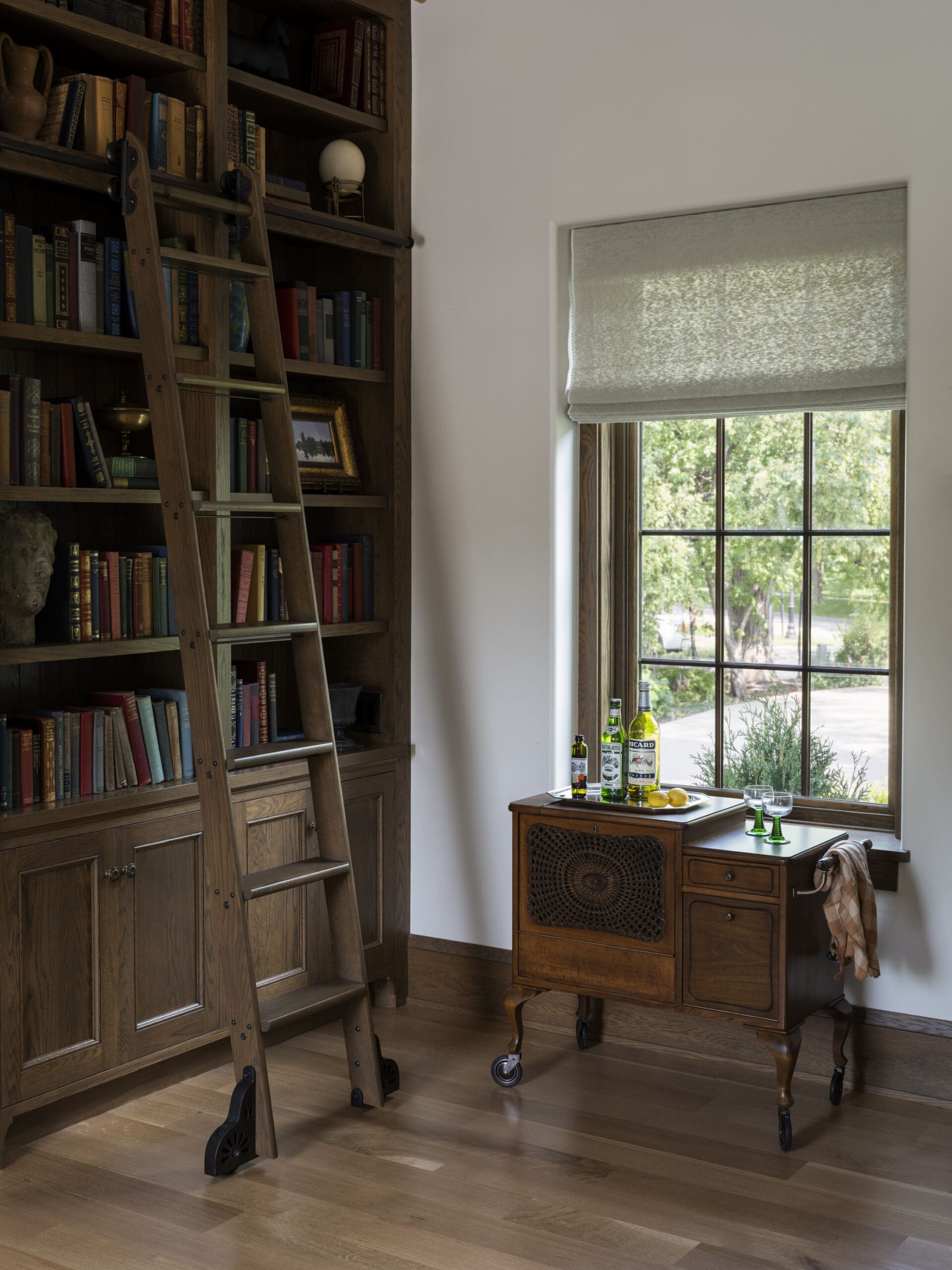 Wood-paneled library with a tall bookcase and a wooden ladder, near a window with a shade; a rolling cart on wheels holds liquor bottles and glassware.