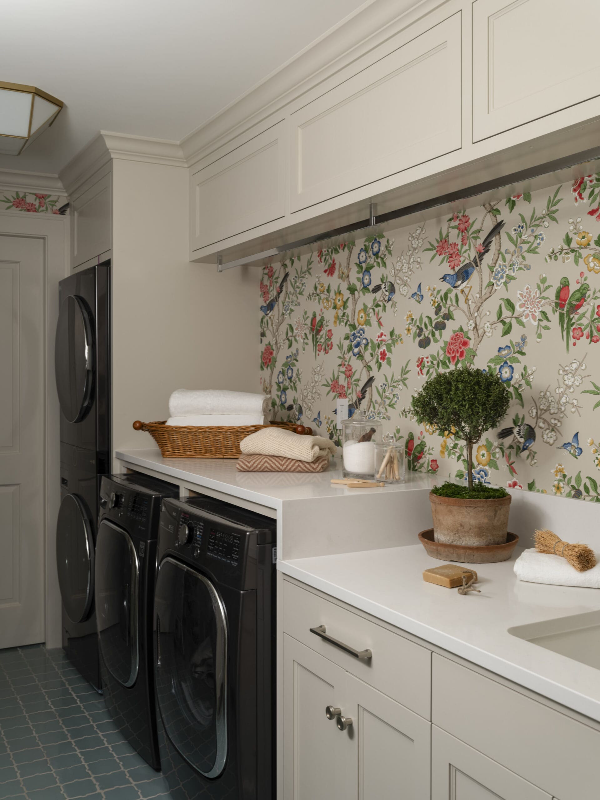 Laundry room with white cabinets above a counter, front-loading washer and dryer, and a basket of towels on the counter beneath floral wallpaper.