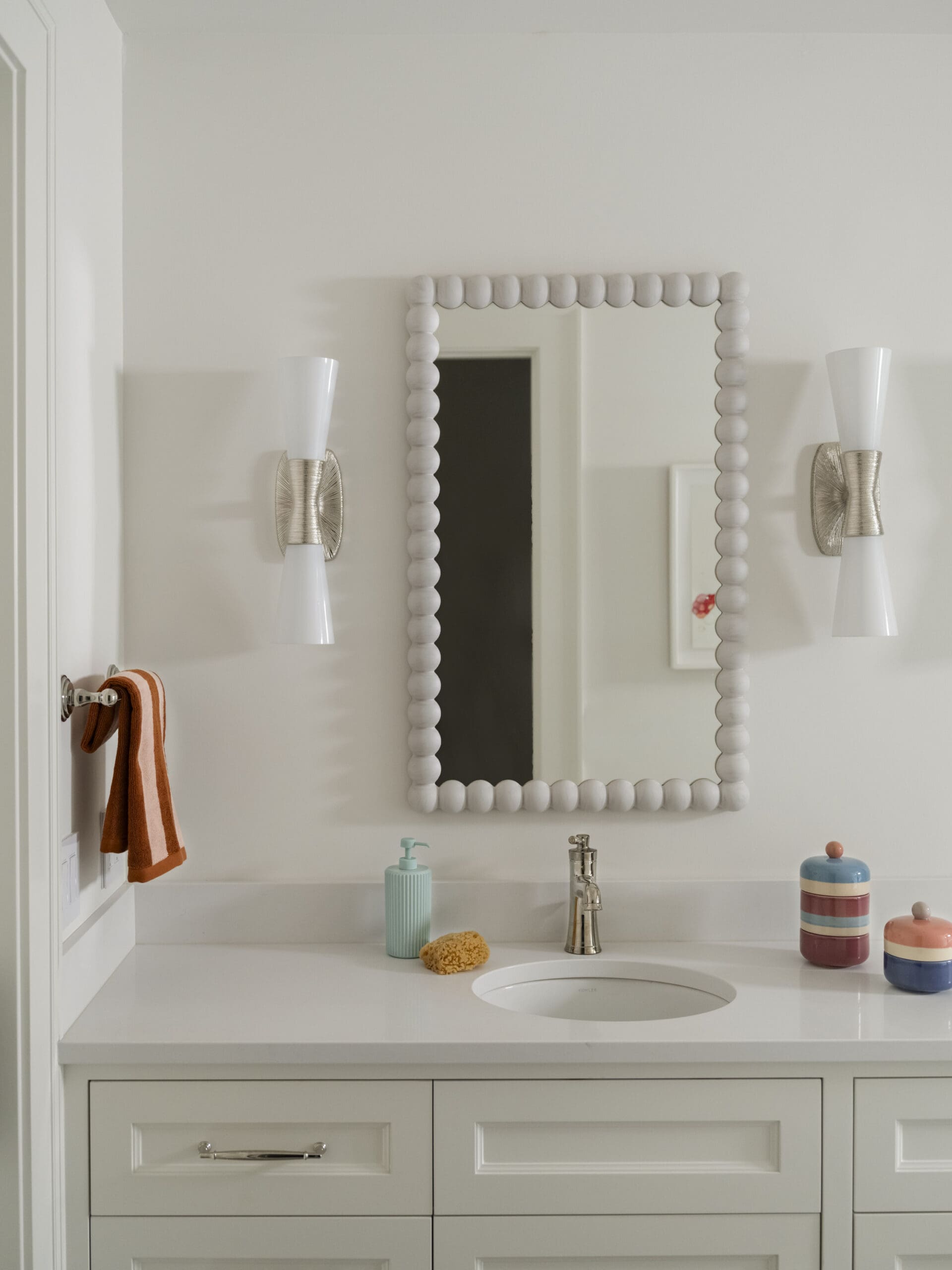 White bathroom vanity with a beaded square mirror and two white wall sconces on either side, orange towel hanging left.