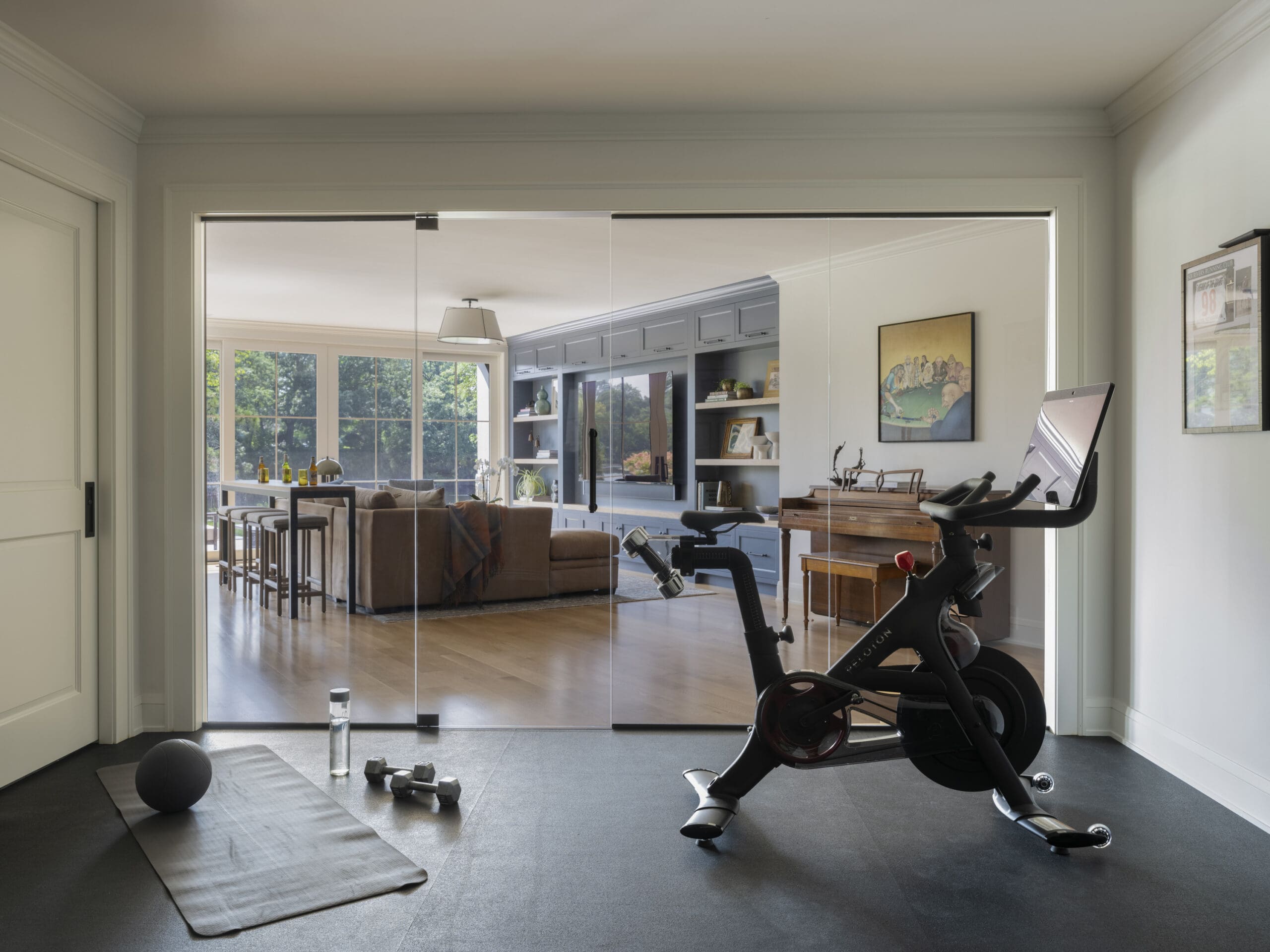 Home gym with two exercise bikes in the foreground, a yoga mat and dumbbells on the floor, and a bright living area visible through glass doors.