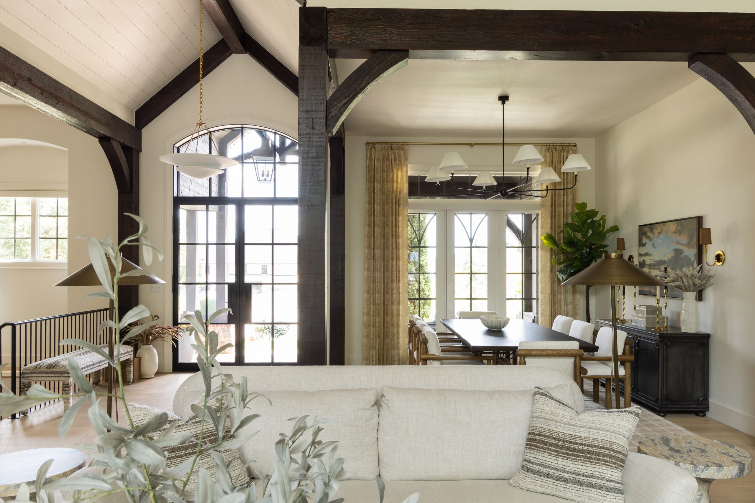 Open-plan living and dining room with dark wood beams, arched windows, and a large chandelier.