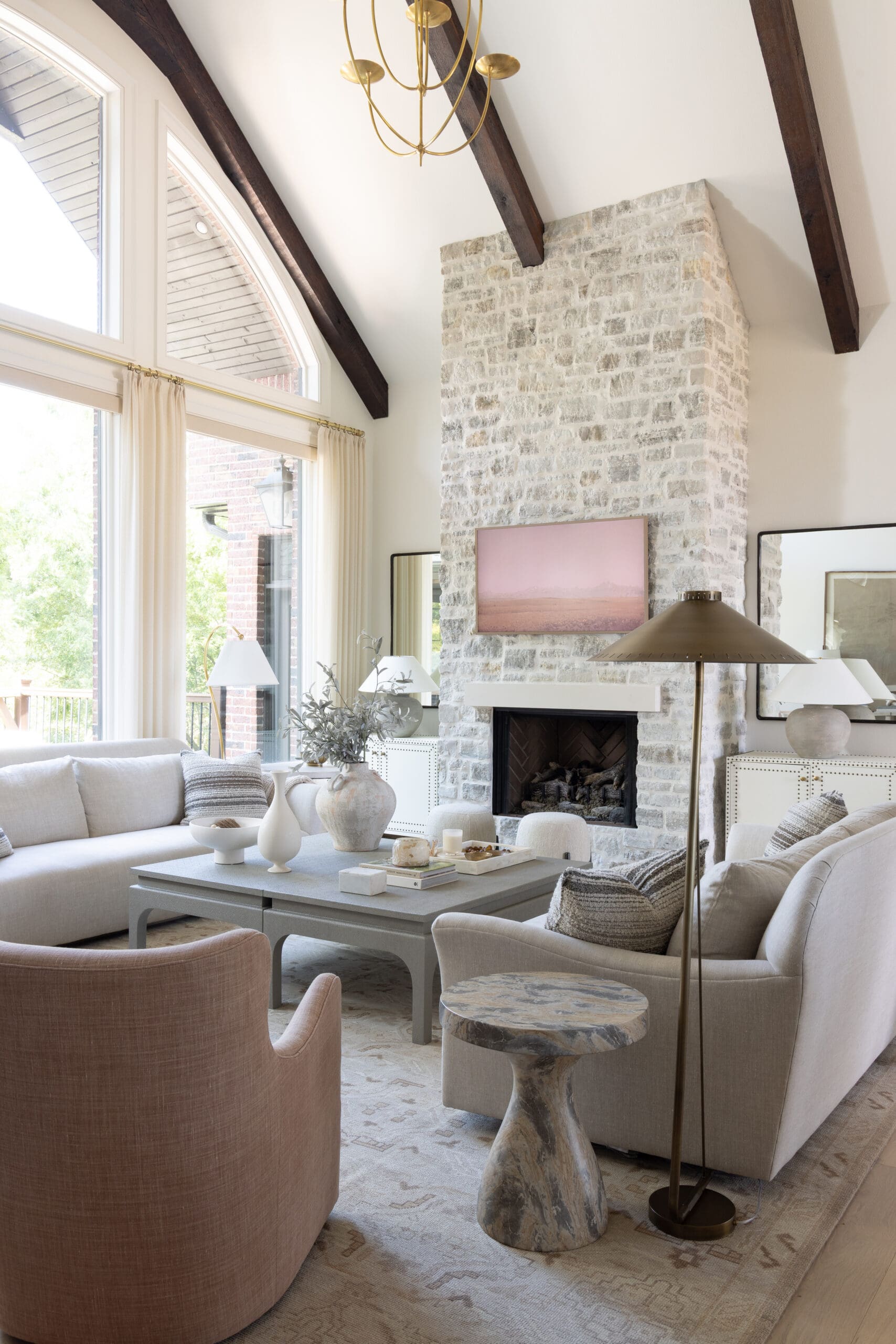 Sunlit living room with a tall stone fireplace, dark wood ceiling beams, and large arched windows over a neutral seating area.