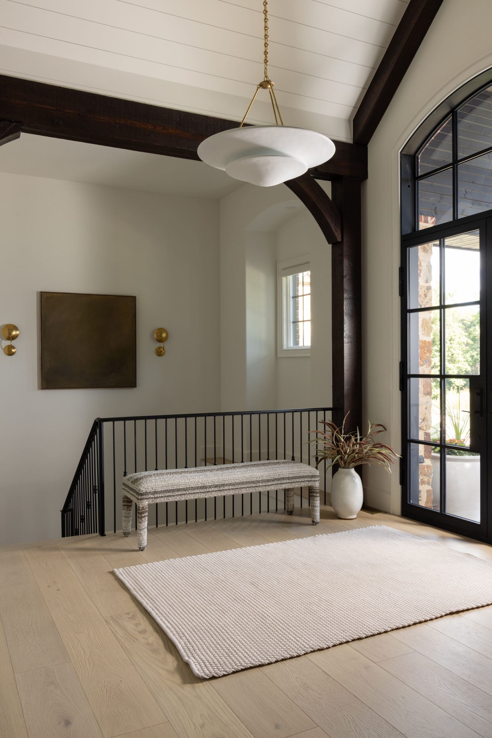 Interior stair landing with a striped upholstered bench, cream rug, black metal railing, and dark wooden beams near a tall arched glass door.",