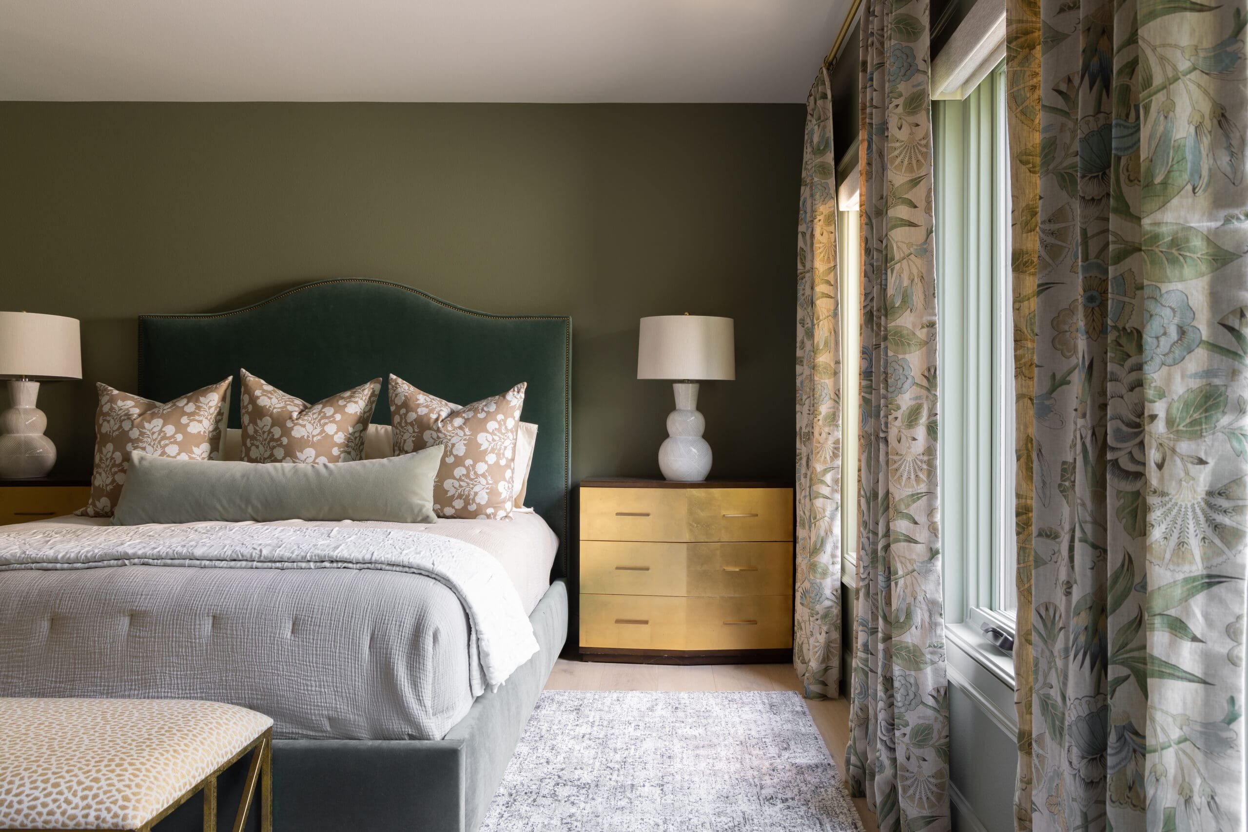 Hotel-style bedroom with dark green velvet headboard, beige floral pillows, and gold bedside dresser beside large floral curtains.