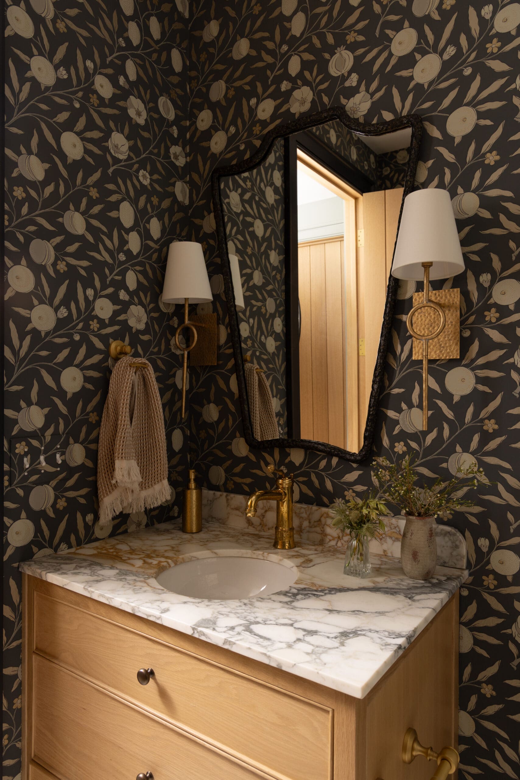 Bathroom vanity with marble countertop, gold faucet, and wooden drawers beneath a dark botanical wallpaper. A large ornate mirror and wall sconces reflect warm light.