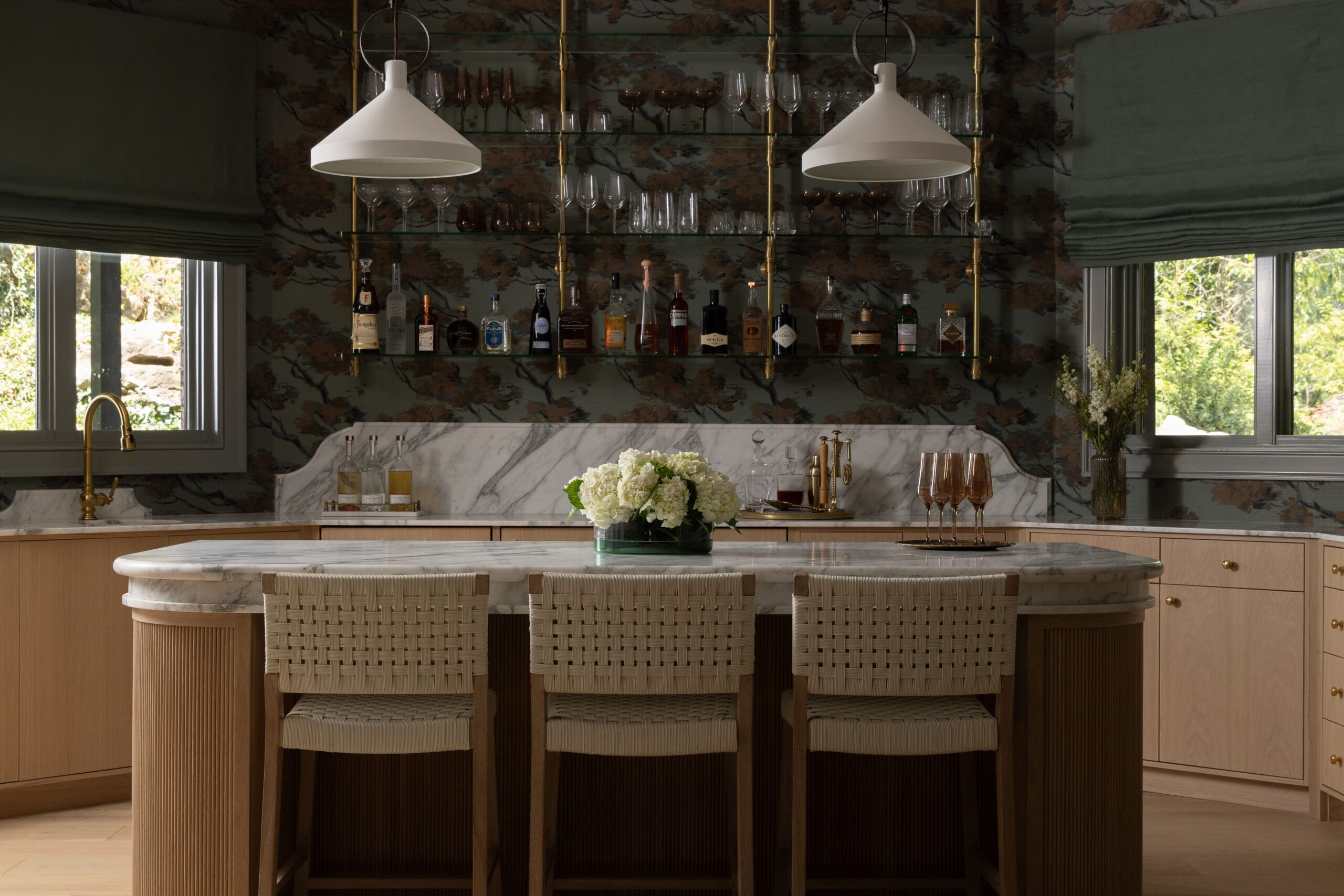 Elegant home bar with a marble countertop, wall-mounted glass shelves of bottles and glassware, and two white pendant lights above wicker bar stools.