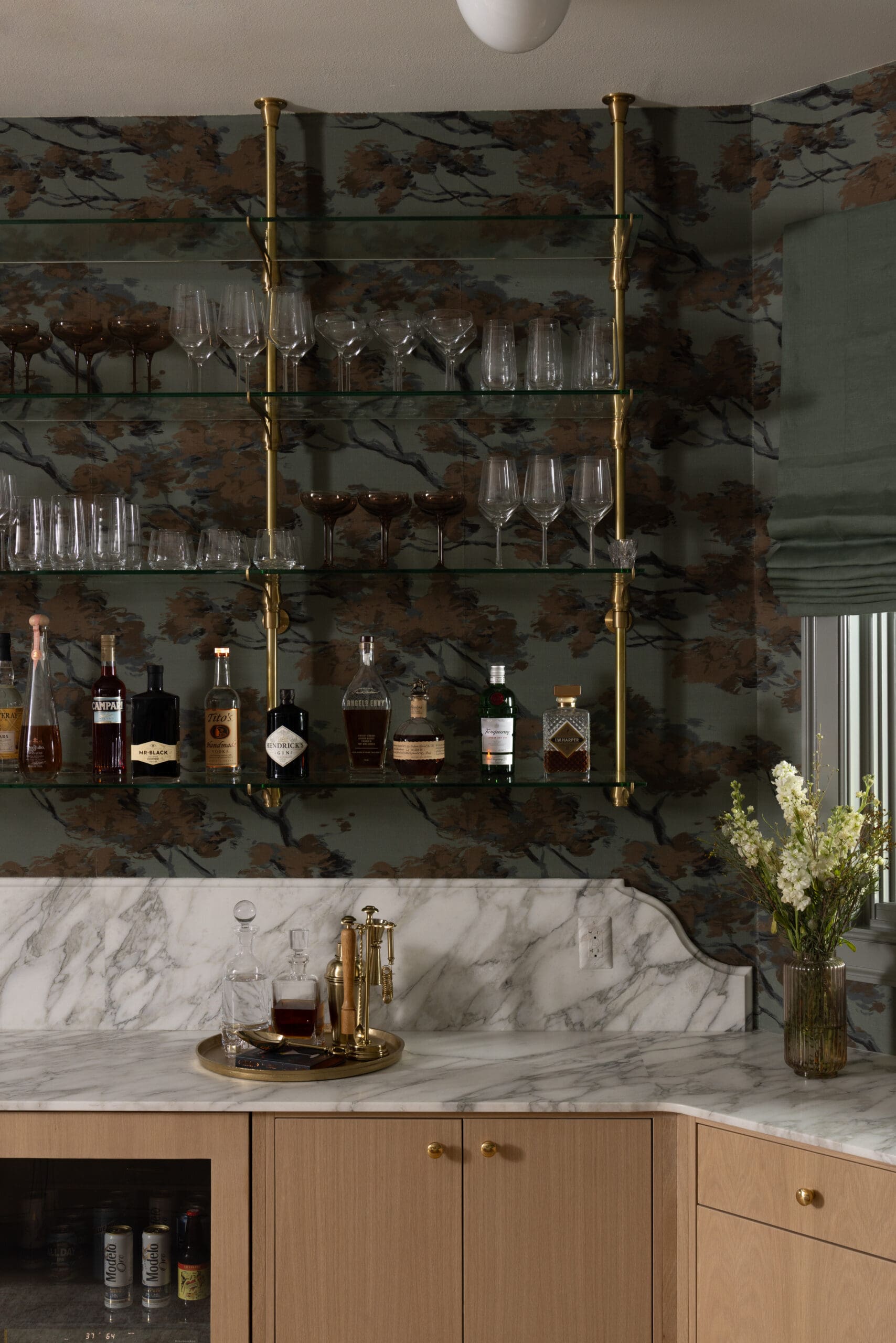 Bar setup with glass shelves on brass supports, holding wine and champagne glasses above a marble counter and liquor bottles below.