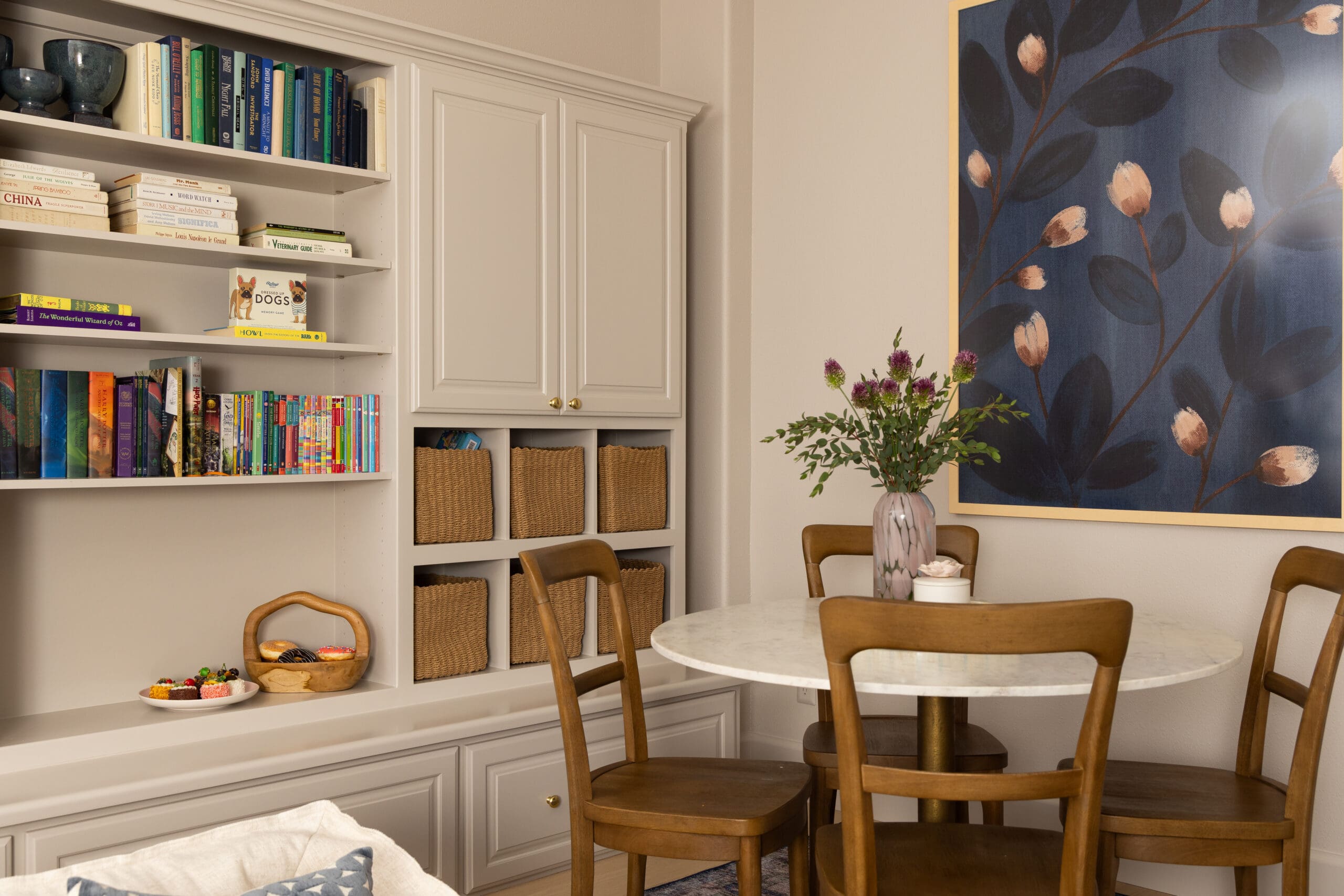 Cozy dining nook with a built-in white bookshelf, woven baskets, a round marble table, and wooden chairs beside blue floral wallpaper.