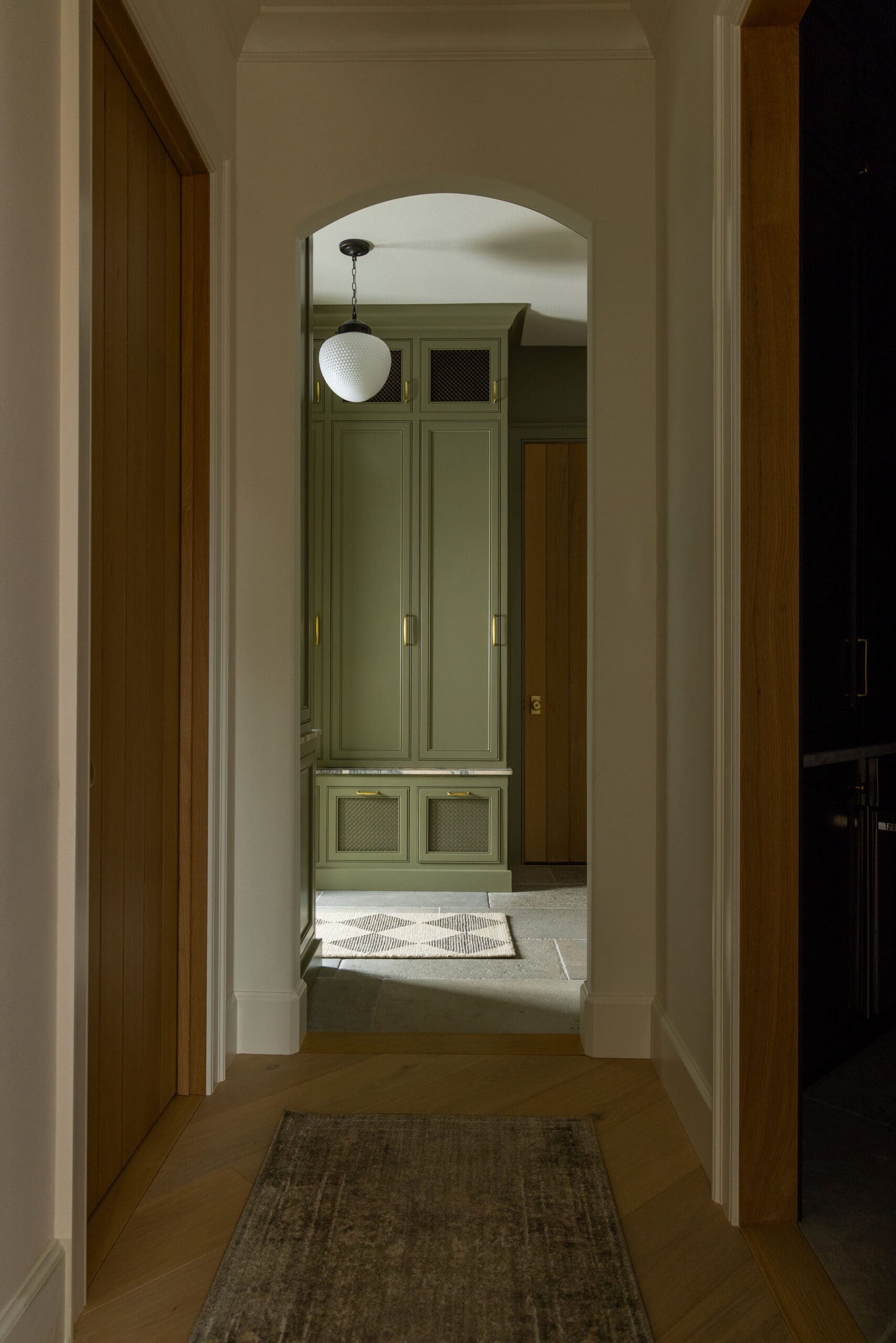 Hallway view through an arched doorway into a kitchen with olive-green cabinets and a white globe pendant light.