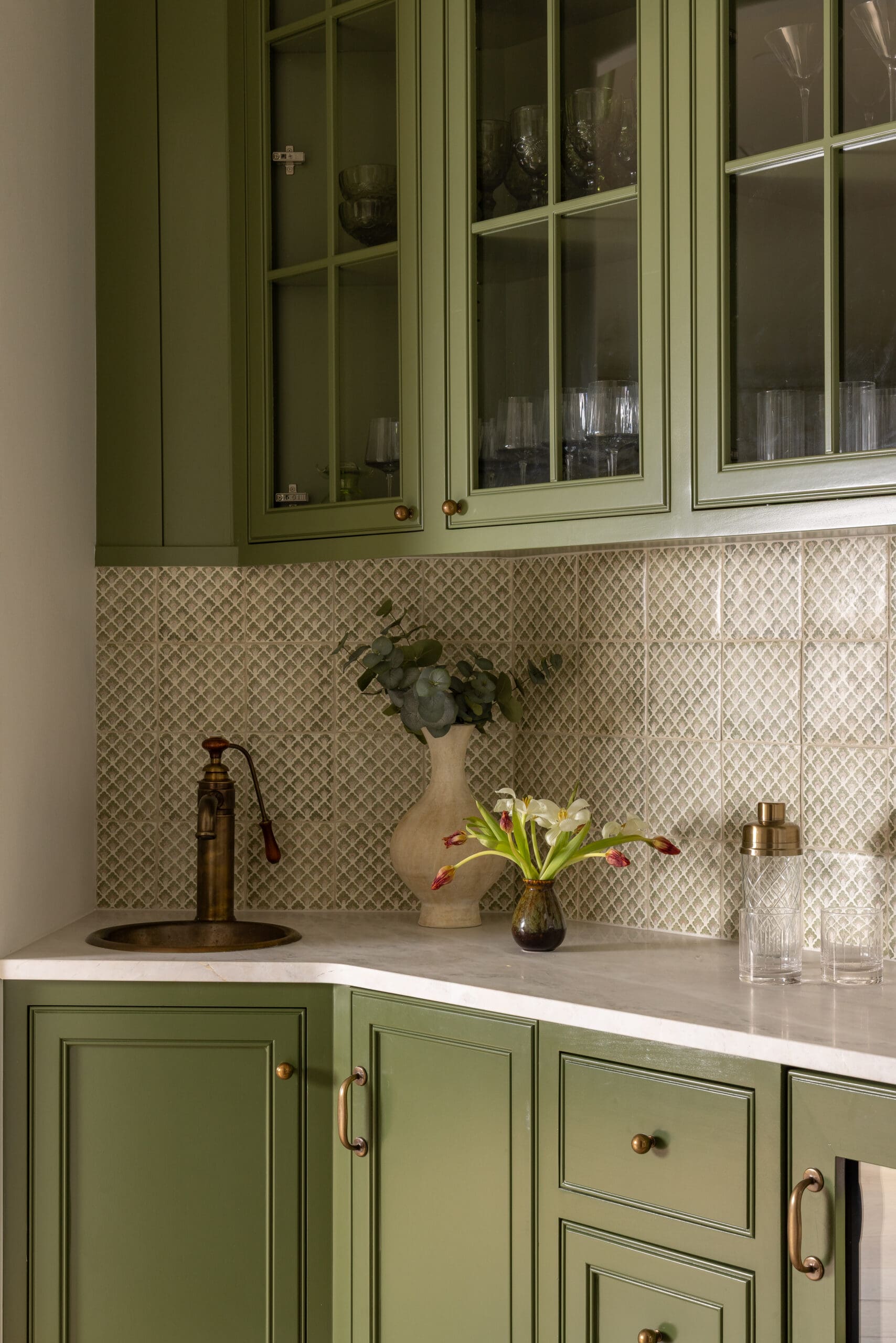 Green kitchen with glass-front upper cabinets, brass knobs, and a vase of flowers on a white marble counter; patterned tile backsplash and glassware inside the cabinet.
