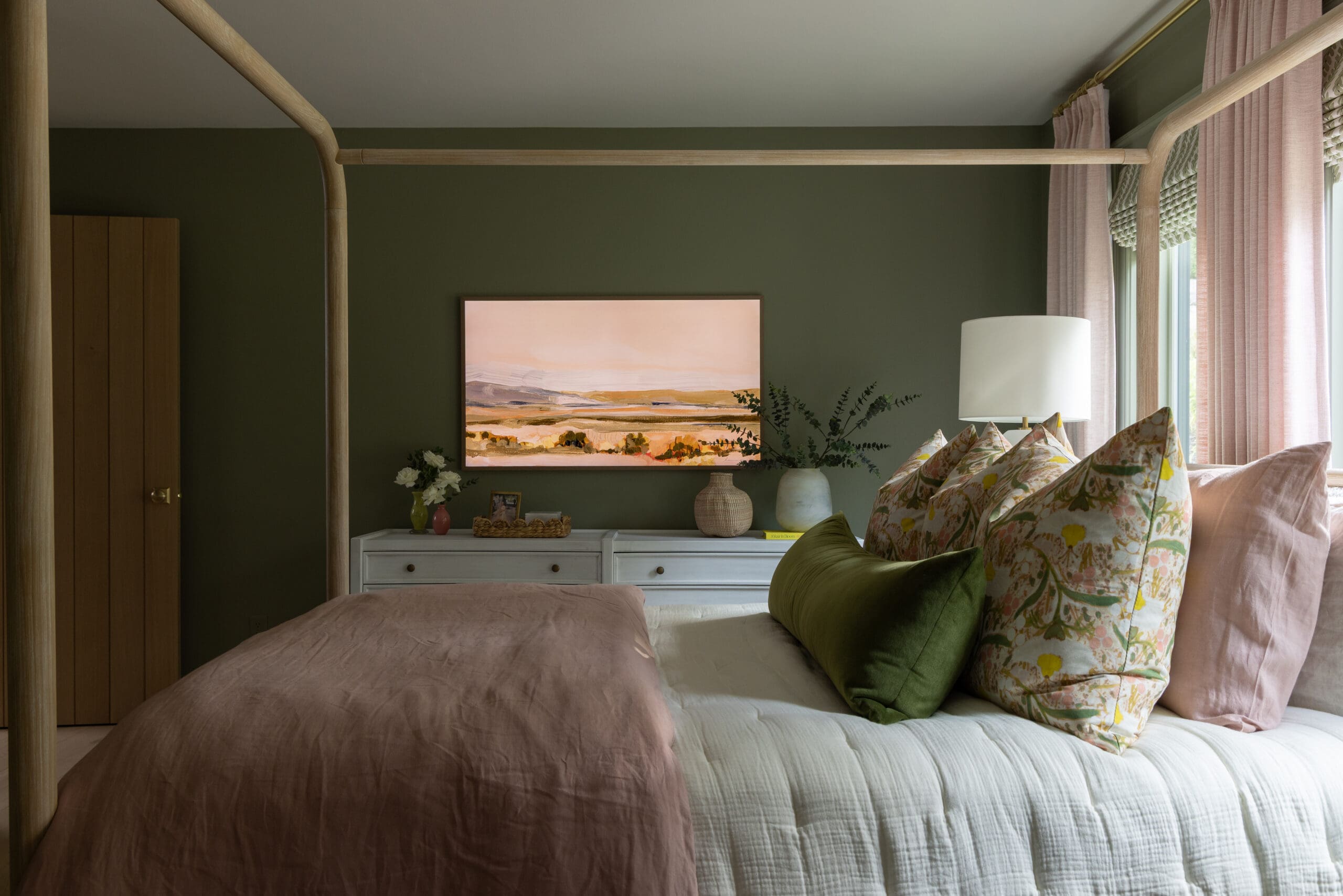 A bedroom scene with a wooden four-poster bed, dusty pink bedding, and floral pillows against a dark green wall. A dresser and landscape painting sit above it.