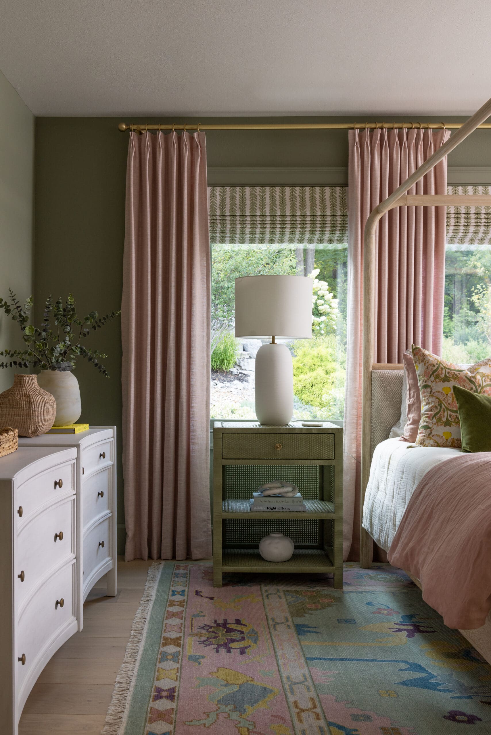 Bedroom with soft pink curtains on brass rod, green walls, and a green nightstand by a window view of greenery.