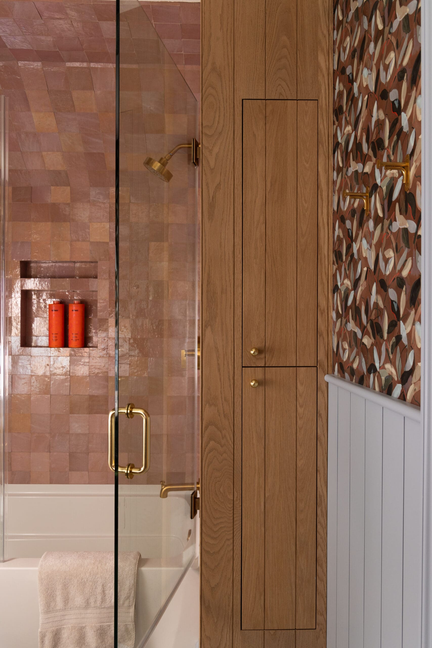 Glass shower with brass handle and pink tile walls, built-in niche with orange bottles, next to a wood-paneled wall and a colorful patterned wallpaper.