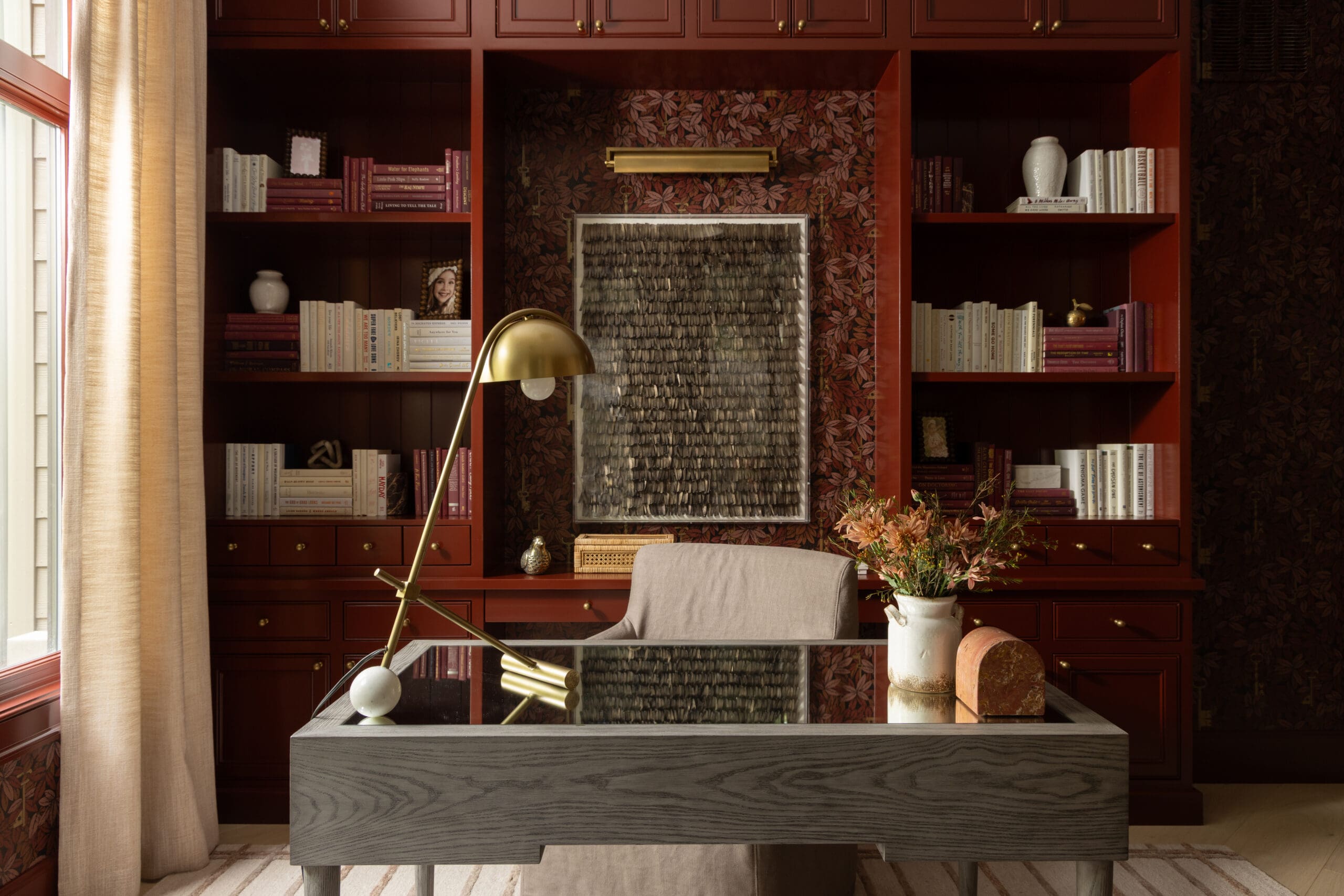 Cozy home office with dark red bookshelves, textured wall art, and a brass desk lamp over a glass-topped desk