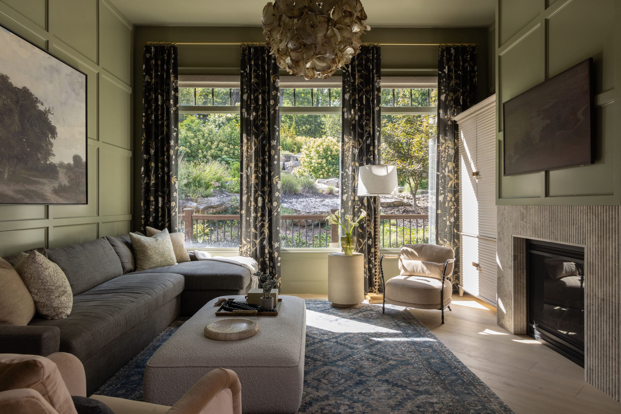 Living room with green paneled walls, a gray sectional sofa, floral curtains, and a shell chandelier; large windows reveal a garden outside.